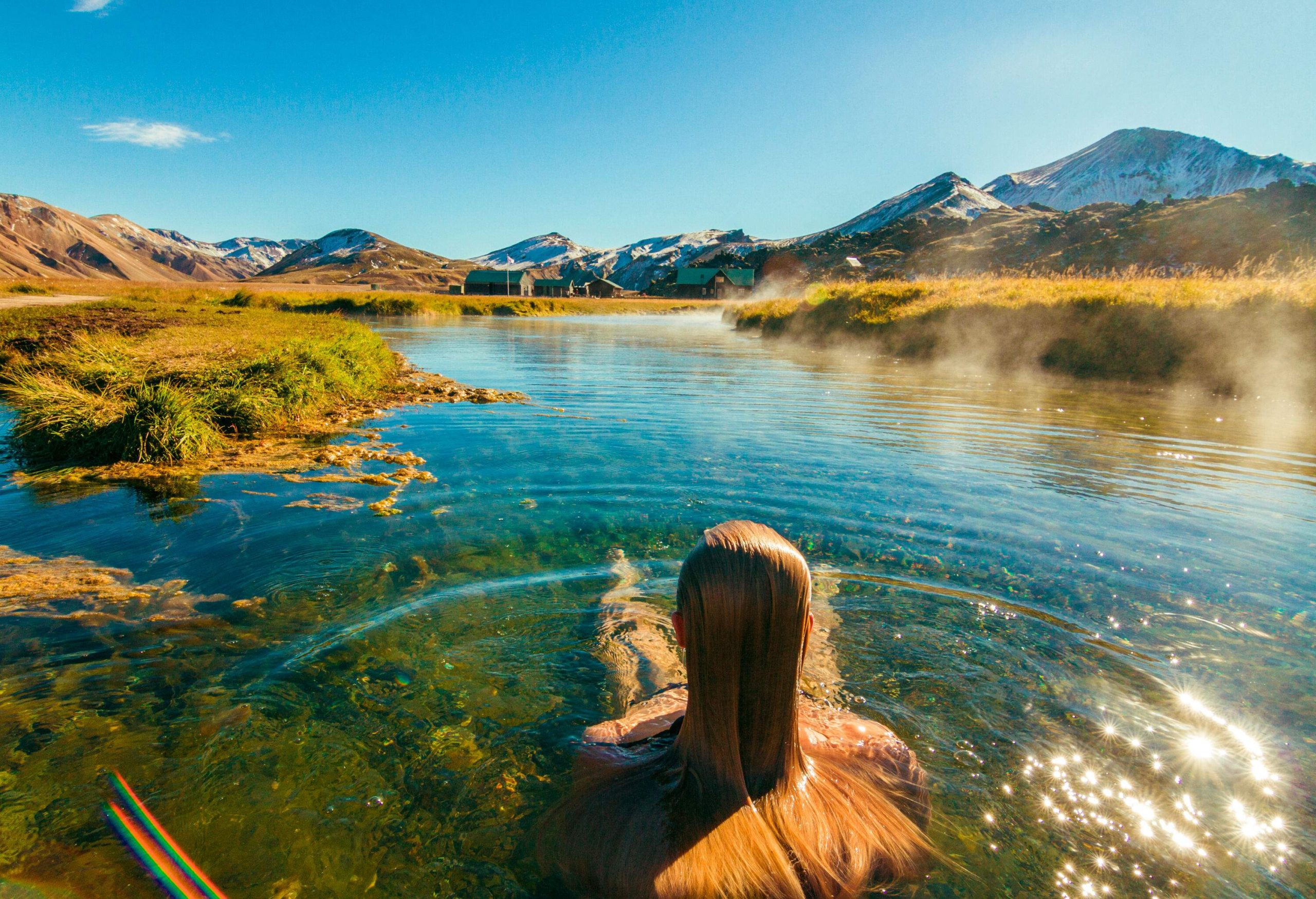 A woman with long blonde hair immersed in a shallow, clear, and steaming hot spring.