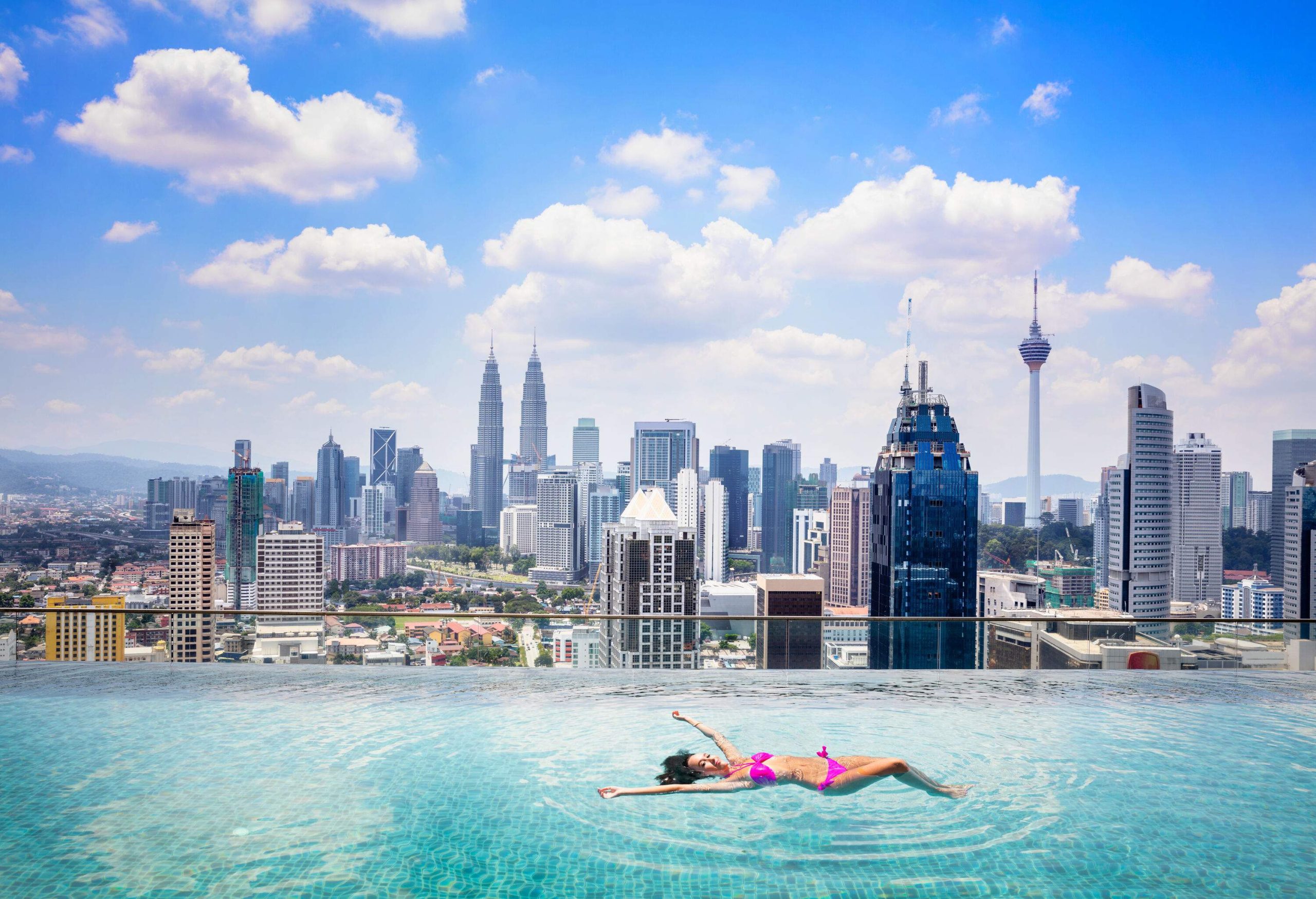 A woman in pink swimwear floats in an infinity swimming pool with a view of modern towering buildings under the cloudy blue sky.