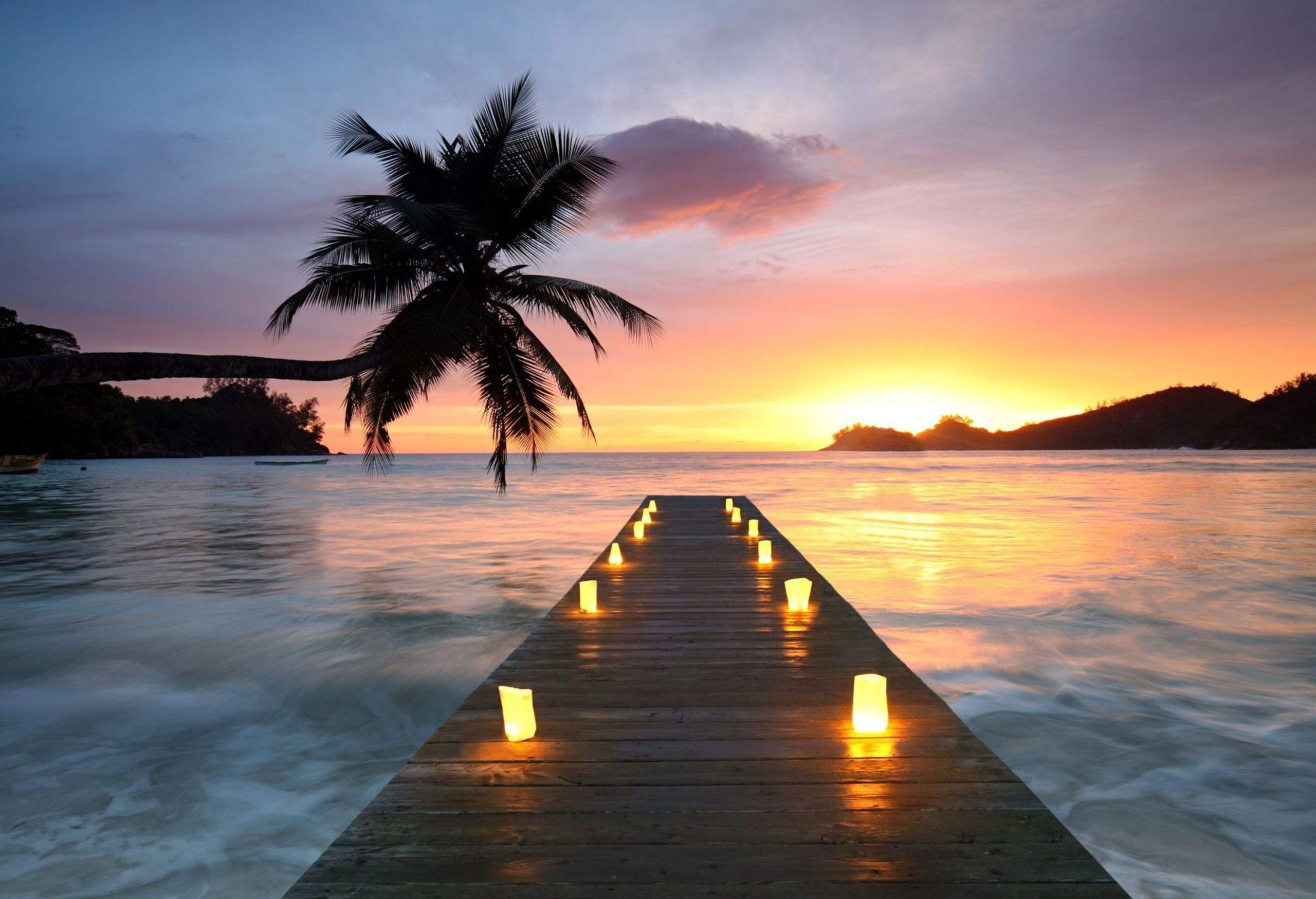 A wooden dock with lit candles extends to the misty seawater with a leaning palm tree on a scenic sunset.