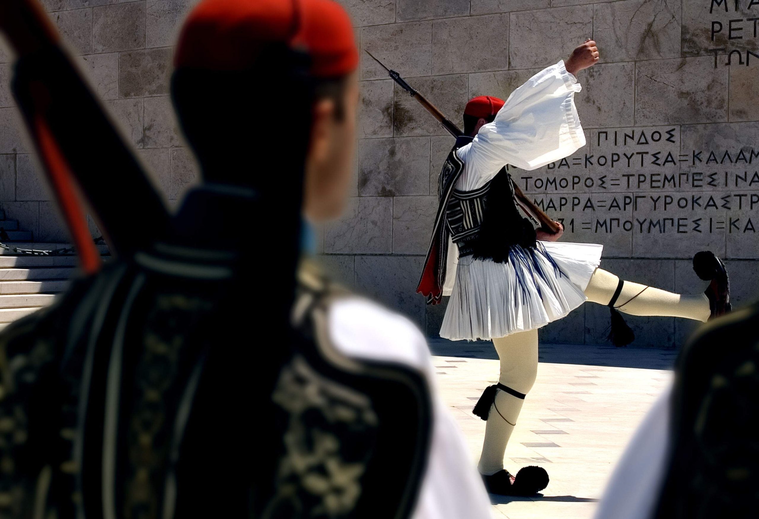 An elite Greek soldier in a traditional Evzones uniform raised his right arm and left leg simultaneously while holding a weapon on his left shoulder.