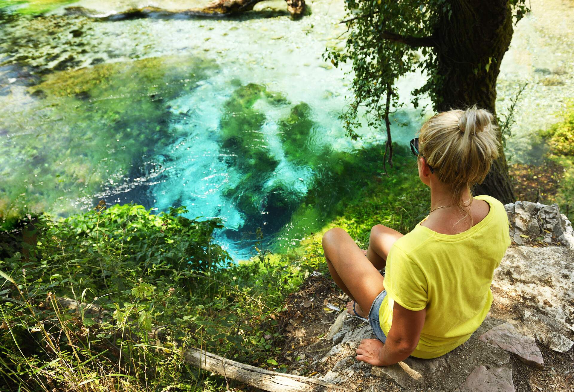 A woman in a yellow shirt sits on a cliff's edge overlooking a spring with turquoise waters.