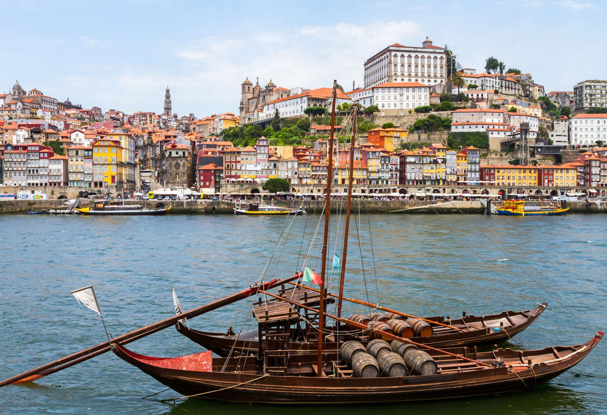 Boats carrying wooden barrels float down the river in the foreground, while other boats sail nearby, with a stunning skyline of towering buildings rising up in the distance.