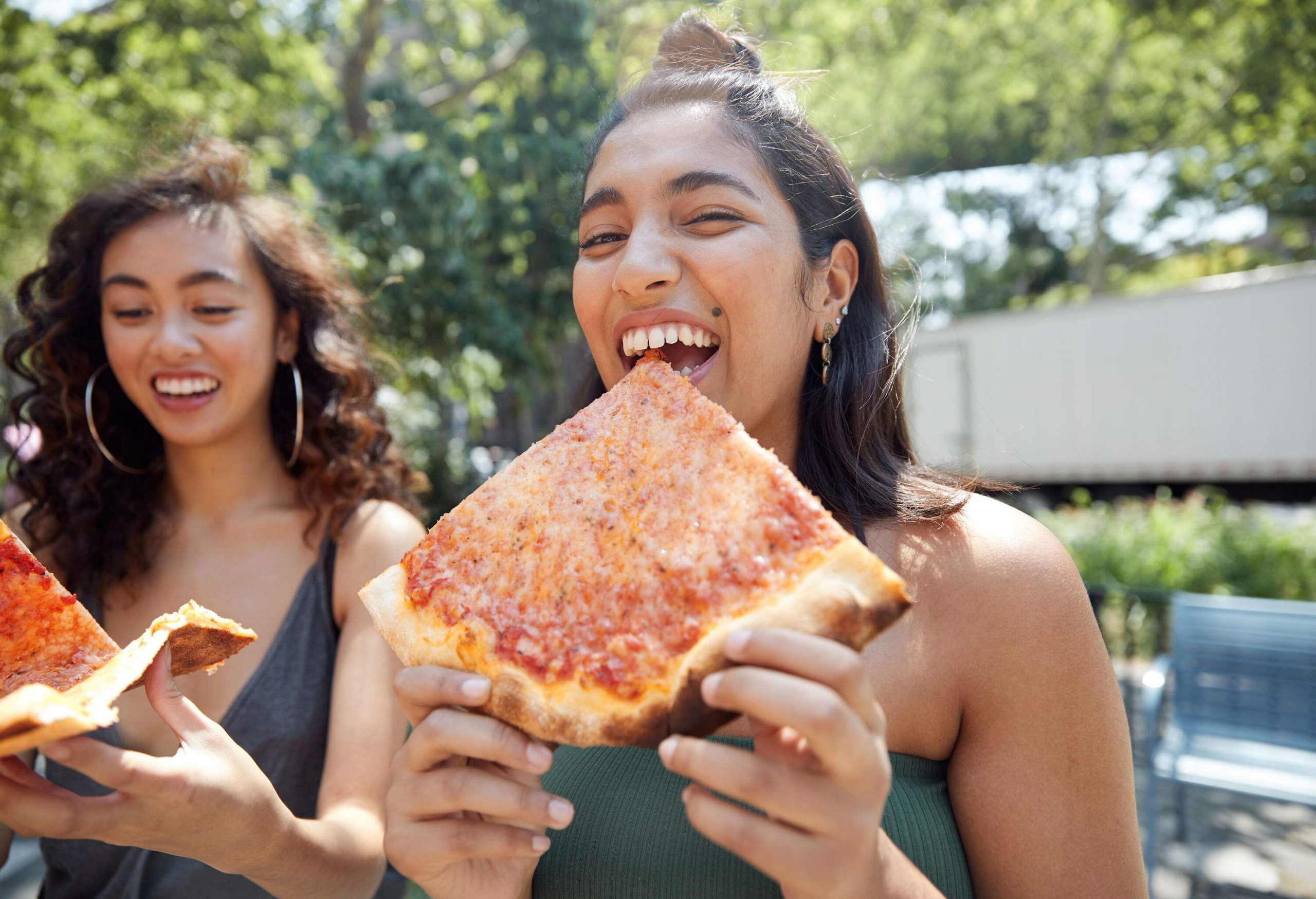 Two young women eating New York–style pizza at a park.