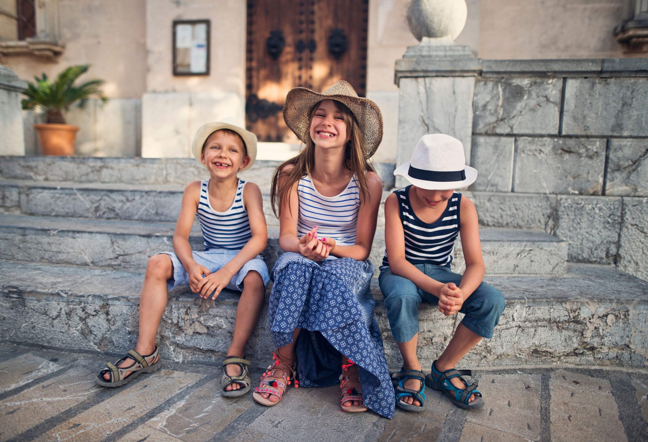 Three children wearing striped sleeveless shirts and summer hats sitting on a staircase.