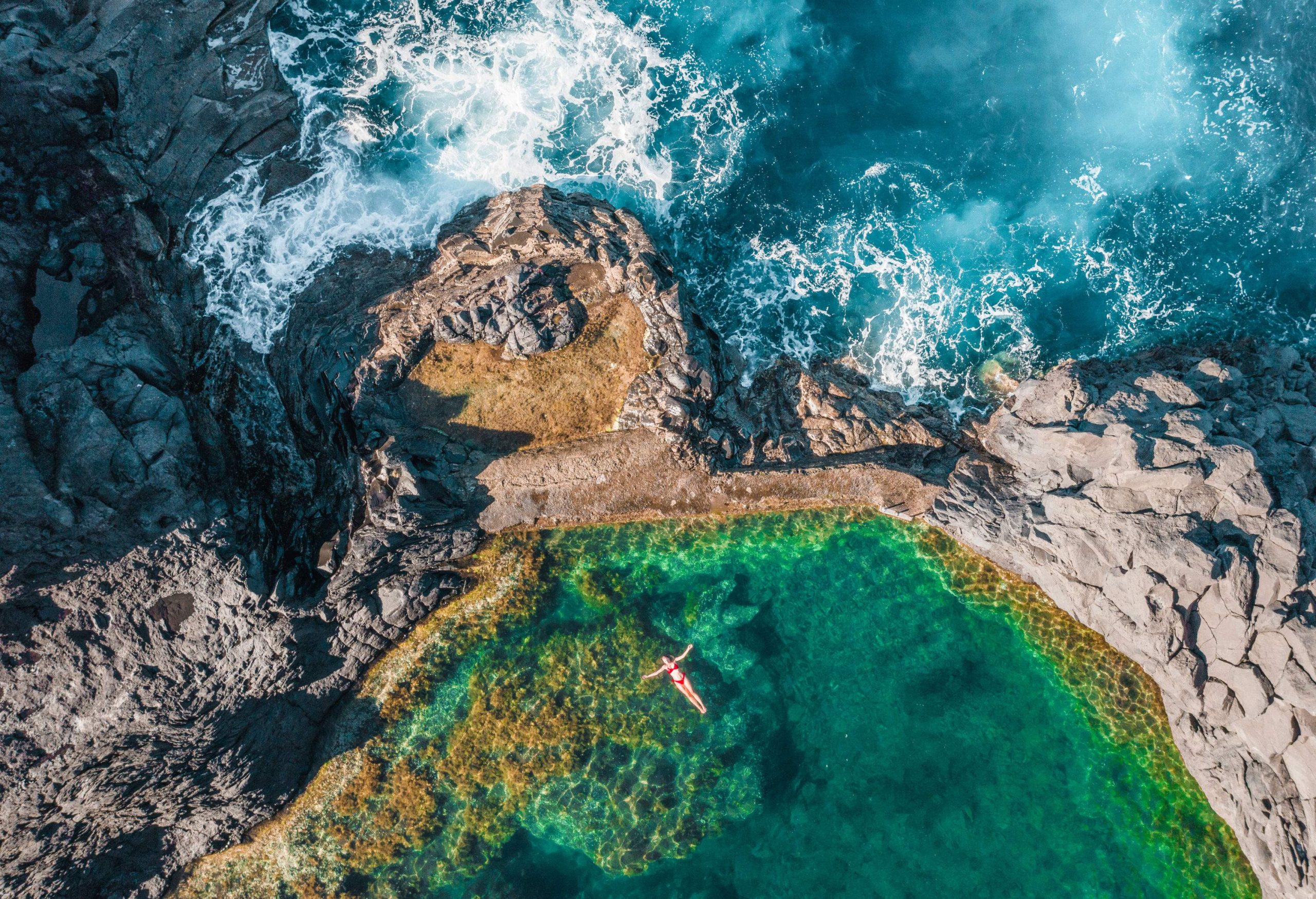 Amidst the rugged beauty of a rocky headland overlooking the coast, a person floats serenely on a pool, with the waves crashing below.