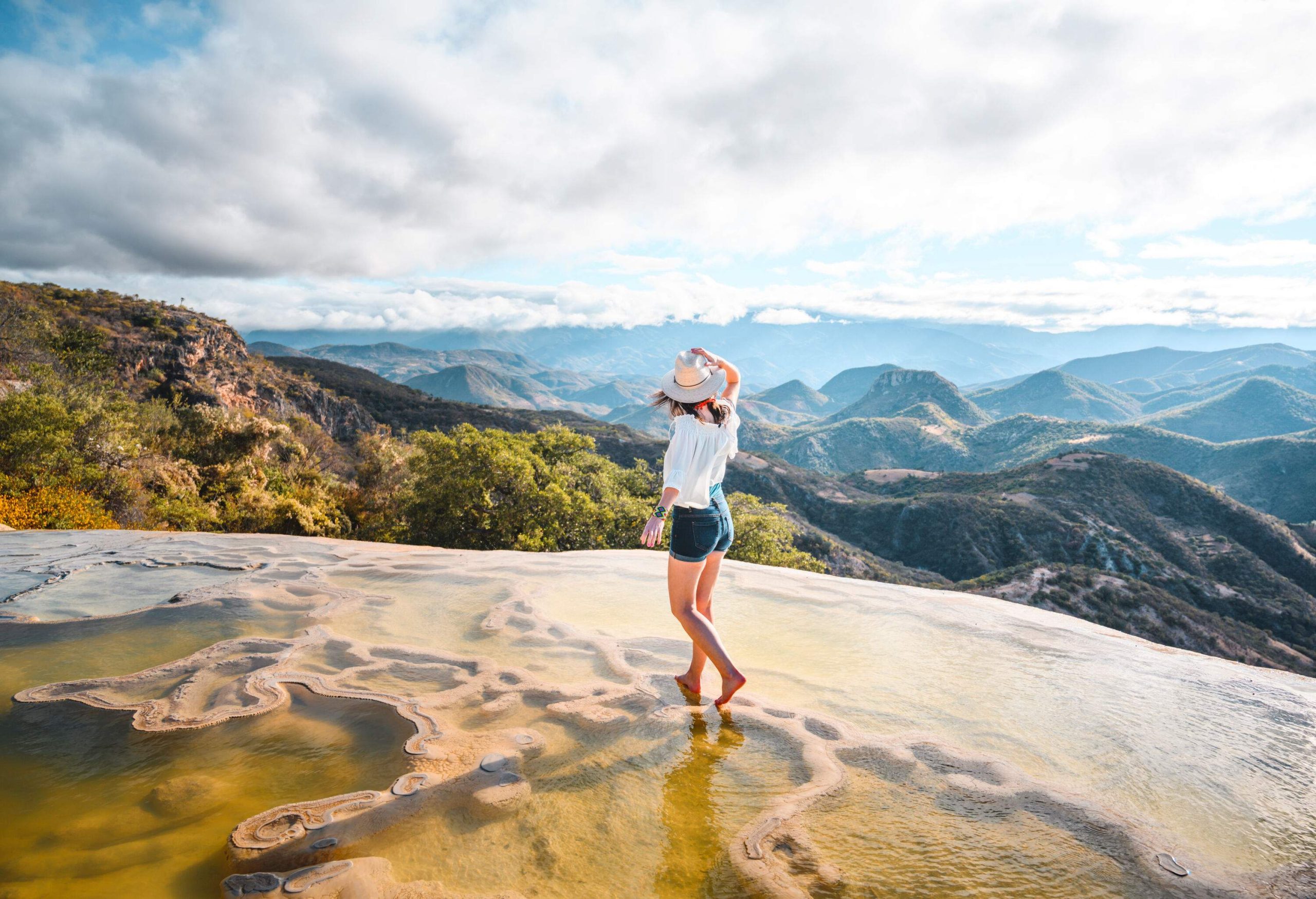A woman stands on the natural rock formations where water trickles over the cliff overlooking the hills and cloud-capped mountains.