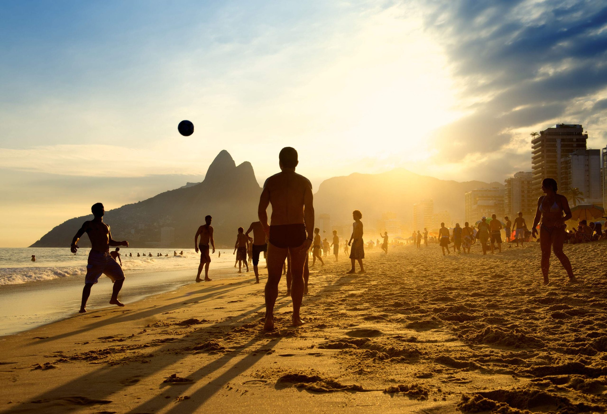 A crowded beach with a group of people playing a ball on a scenic sunset.
