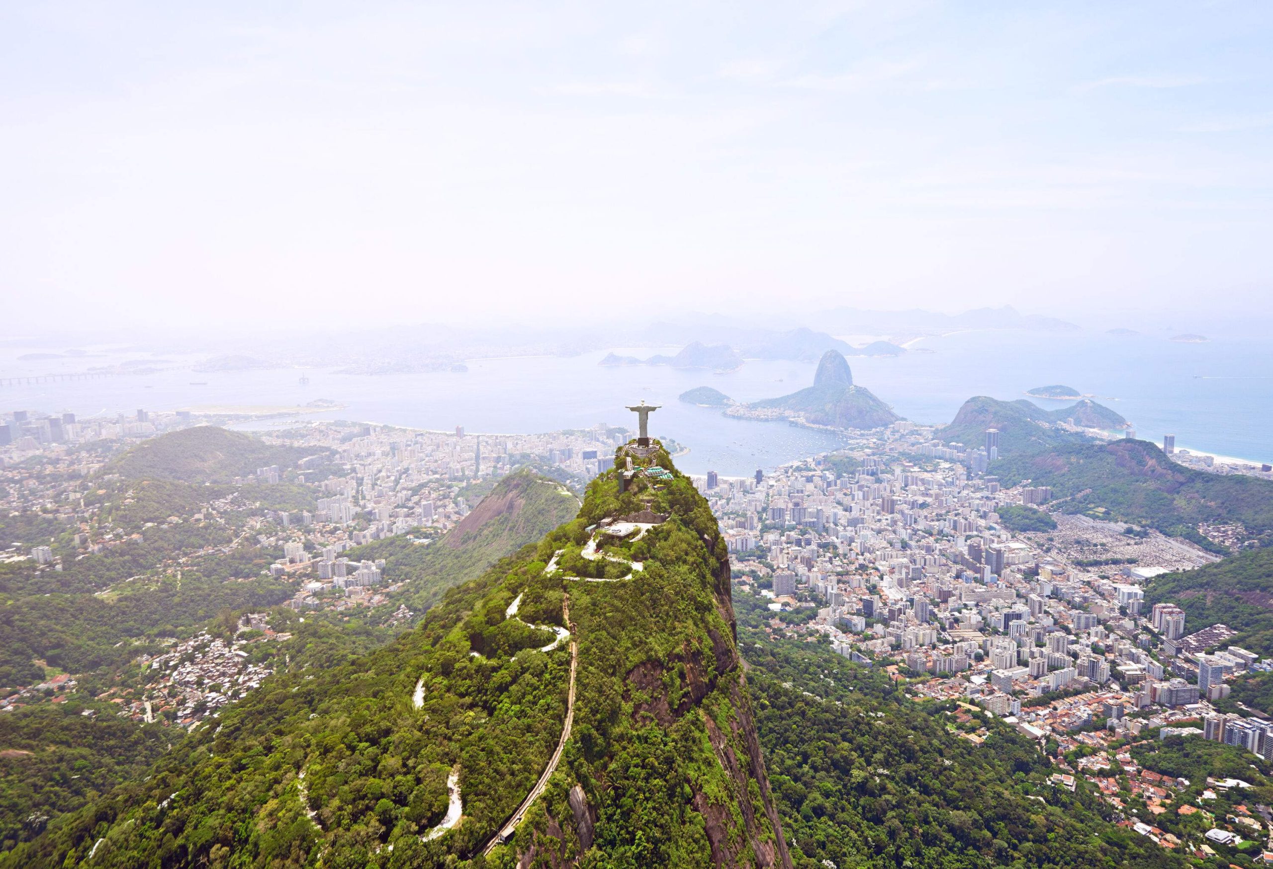 A winding road on a forested hill towards a massive statue of Jesus opening its arms over a cluster of buildings in a coastal city.