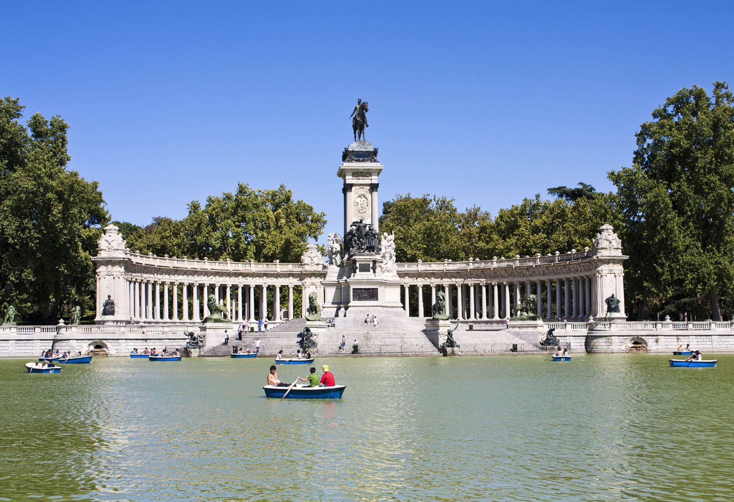 A park's pillared monument with a bronze statue of a person riding a horse overlooking a lake full of people on boats.