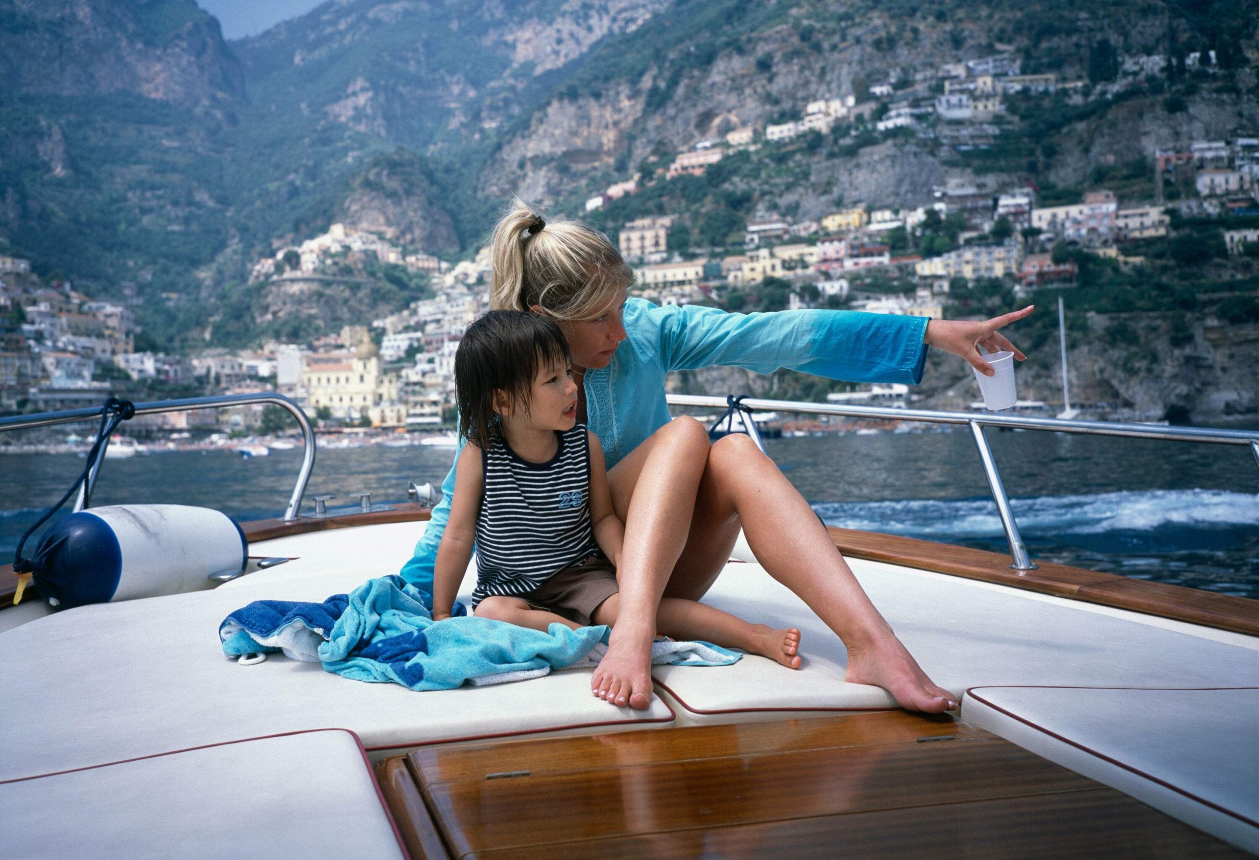 A woman sits beside a kid on a yacht while pointing at the scenery.
