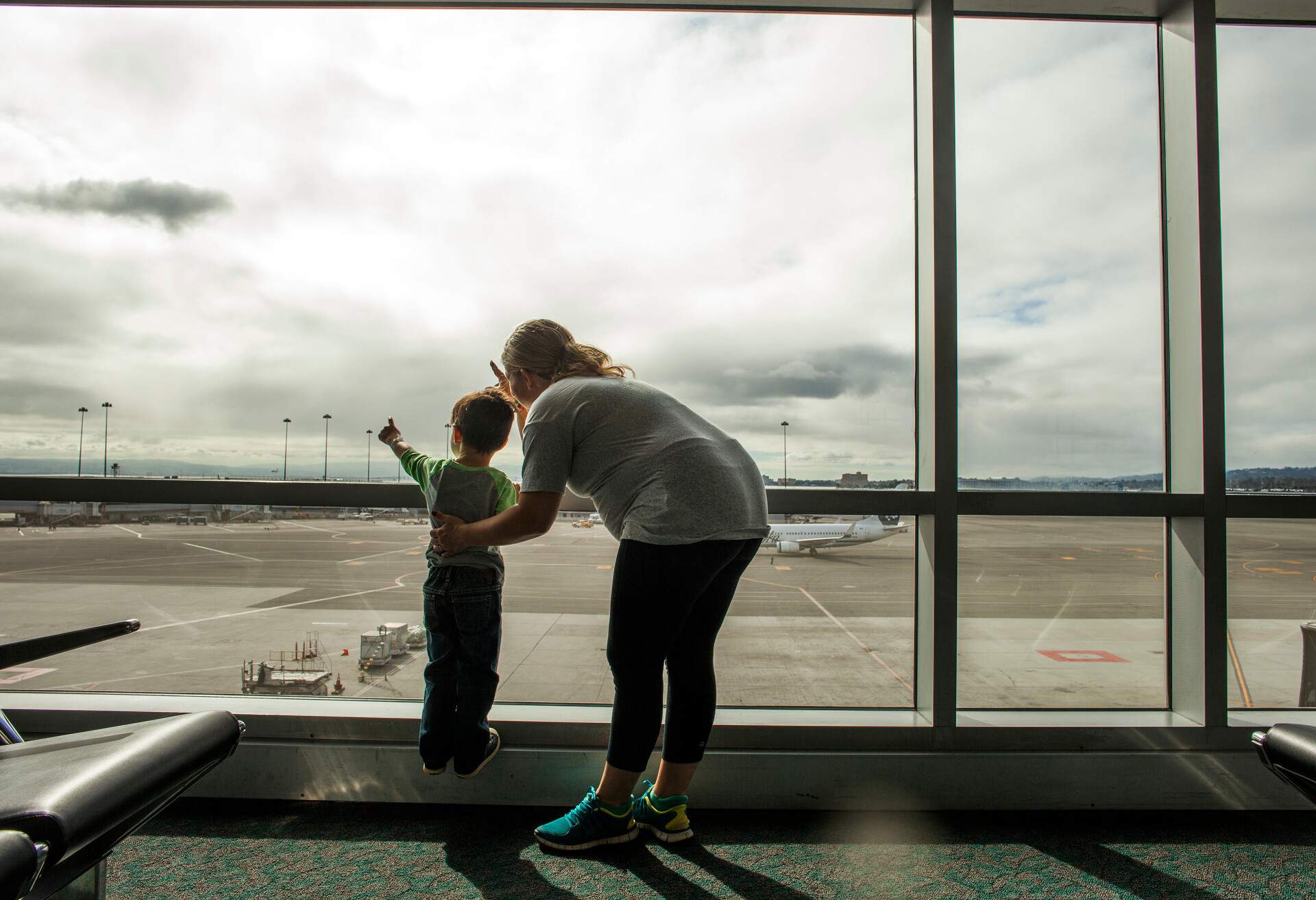 A mother and his son are pointing at the clouds on the airport's glass window.