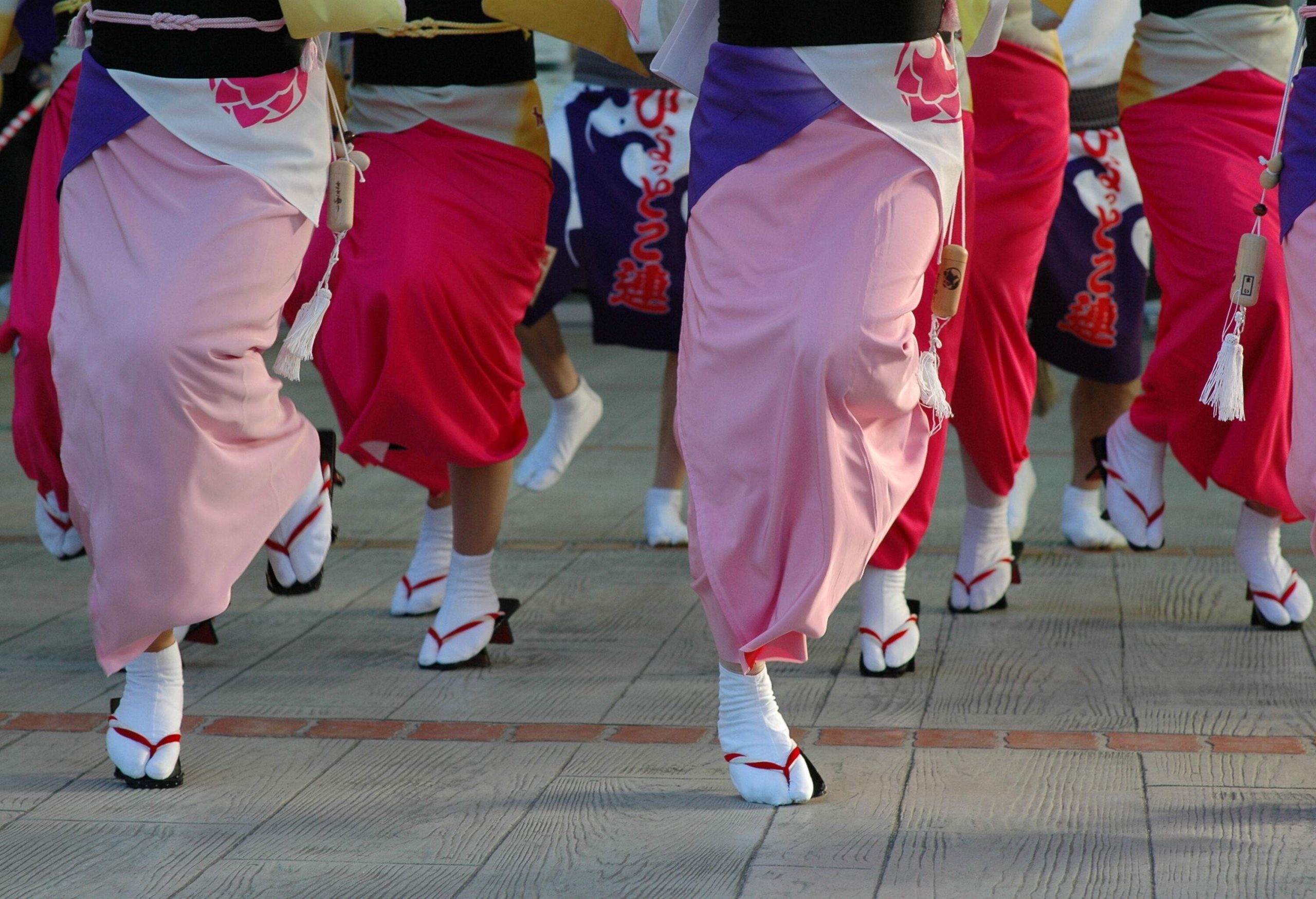 A group of people dance while wearing pink and red sarongs, white socks, and slippers.