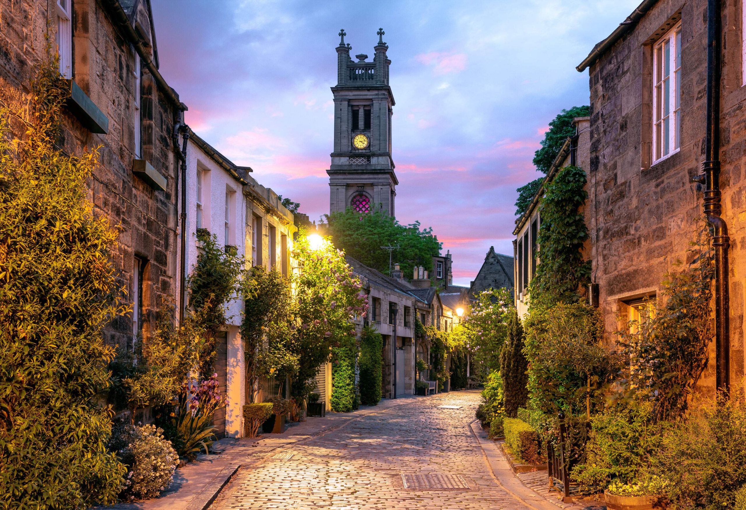 A clock tower looming over a charming cobblestone street lined with plant-covered stone houses.