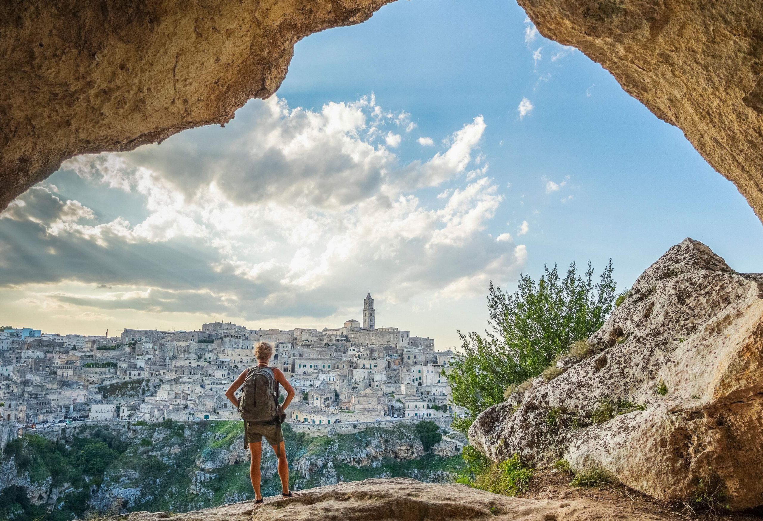 A woman hiker with a backpack stands in the entrance of a cave, surveying the rugged slope and the town perched upon it.