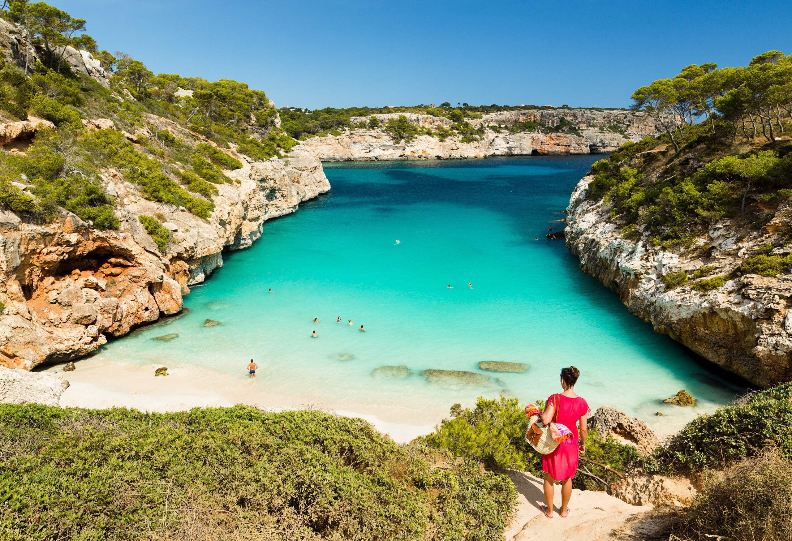 A woman in a pink dress looks at her friends down the beach with white sand and clear waters. 