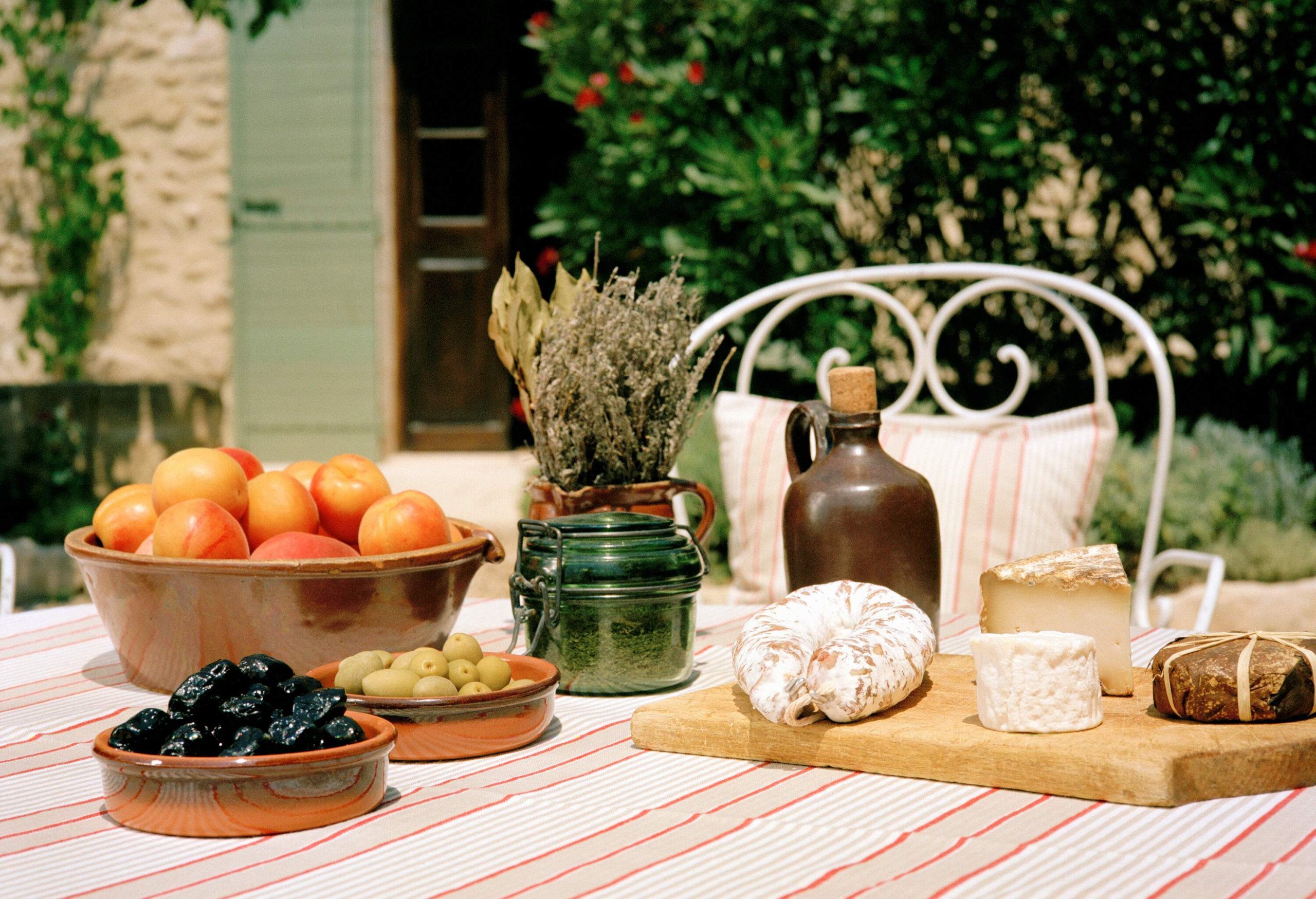 A cloth-covered outdoor table with various fruits, cheese, and a jar.