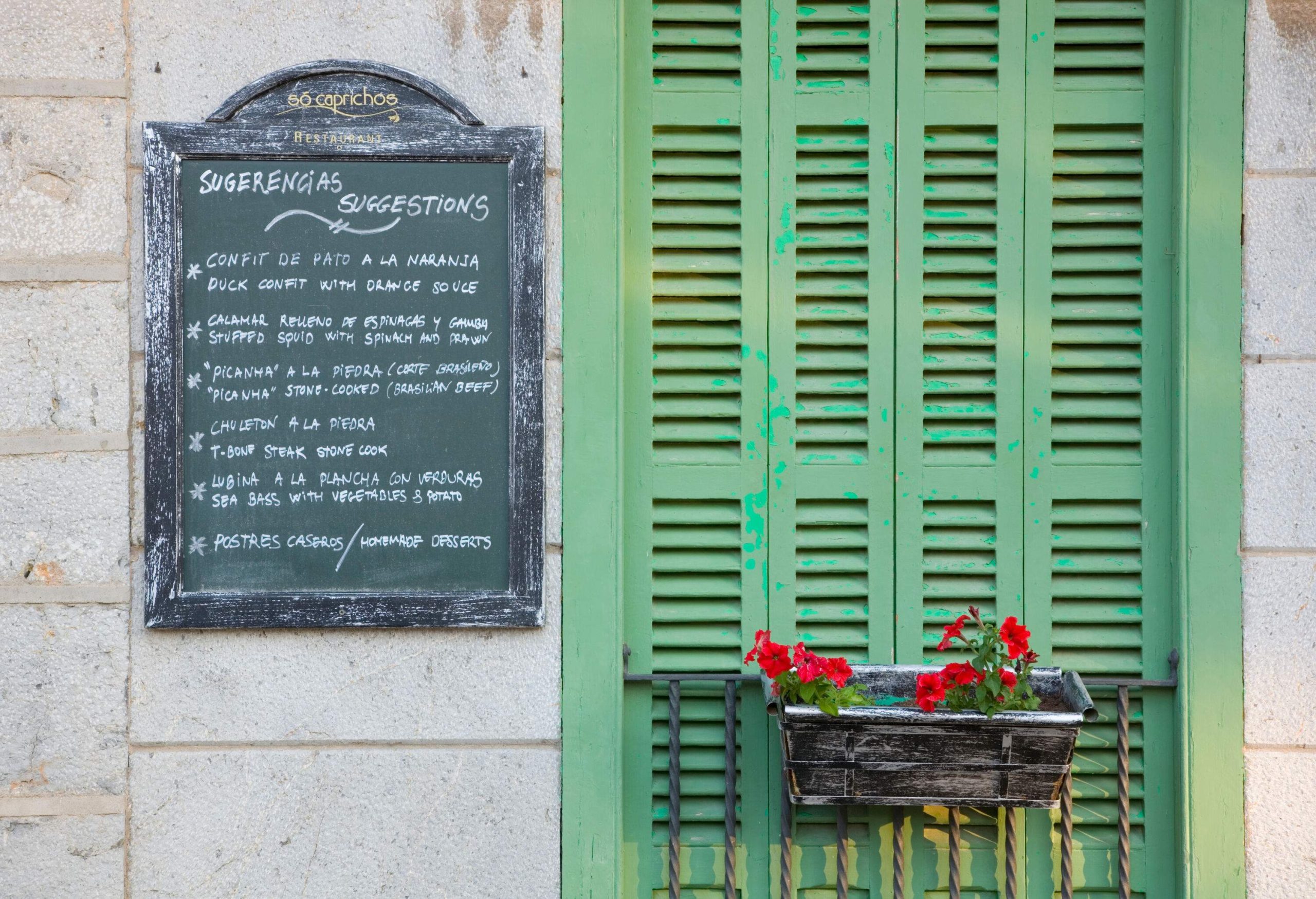 A wooden sign menu board on the wall next to a green window with red flowers on a black plant pot.