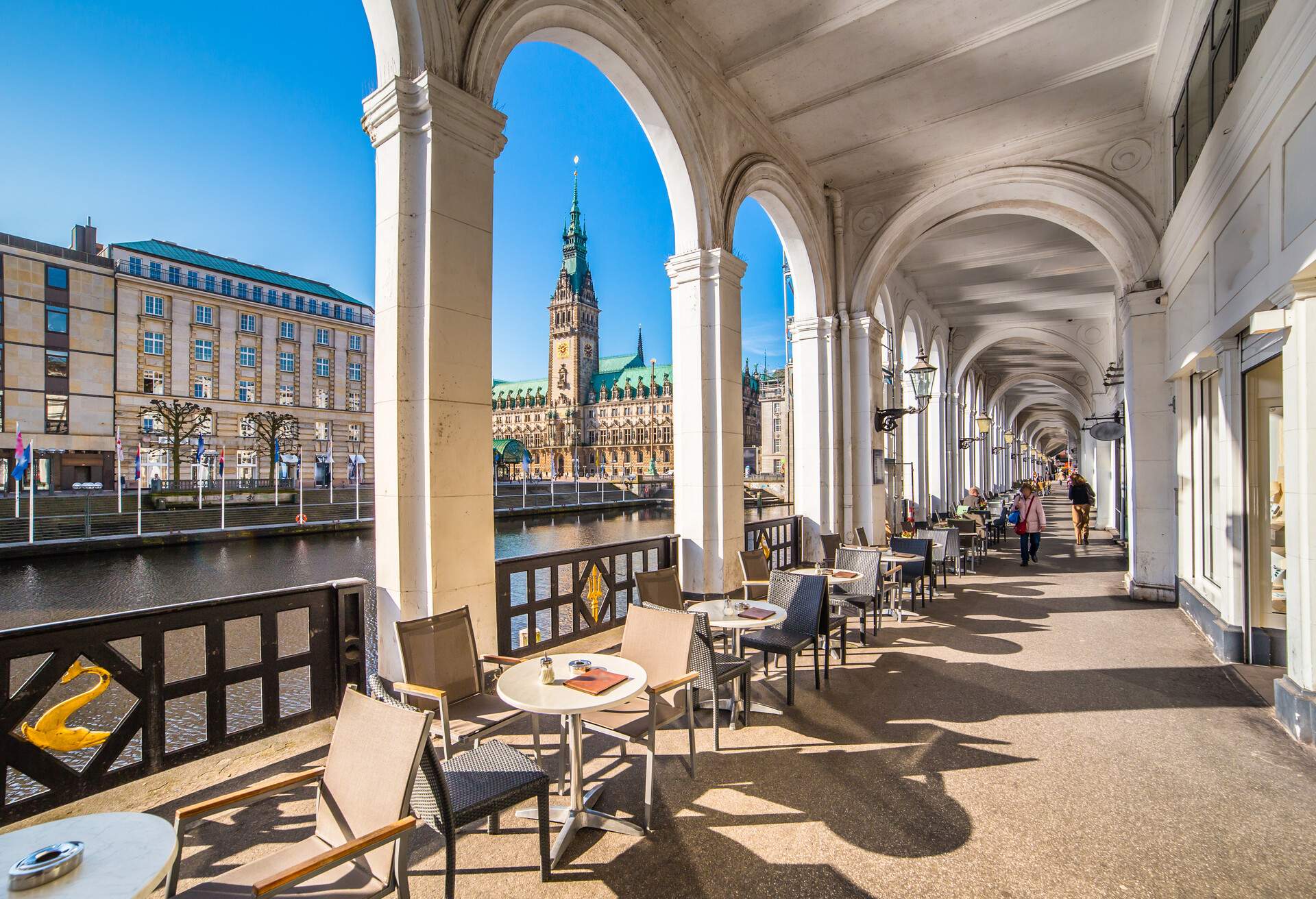 A pillared hallway with chairs and tables overlooking a river canal and waterfront structures.