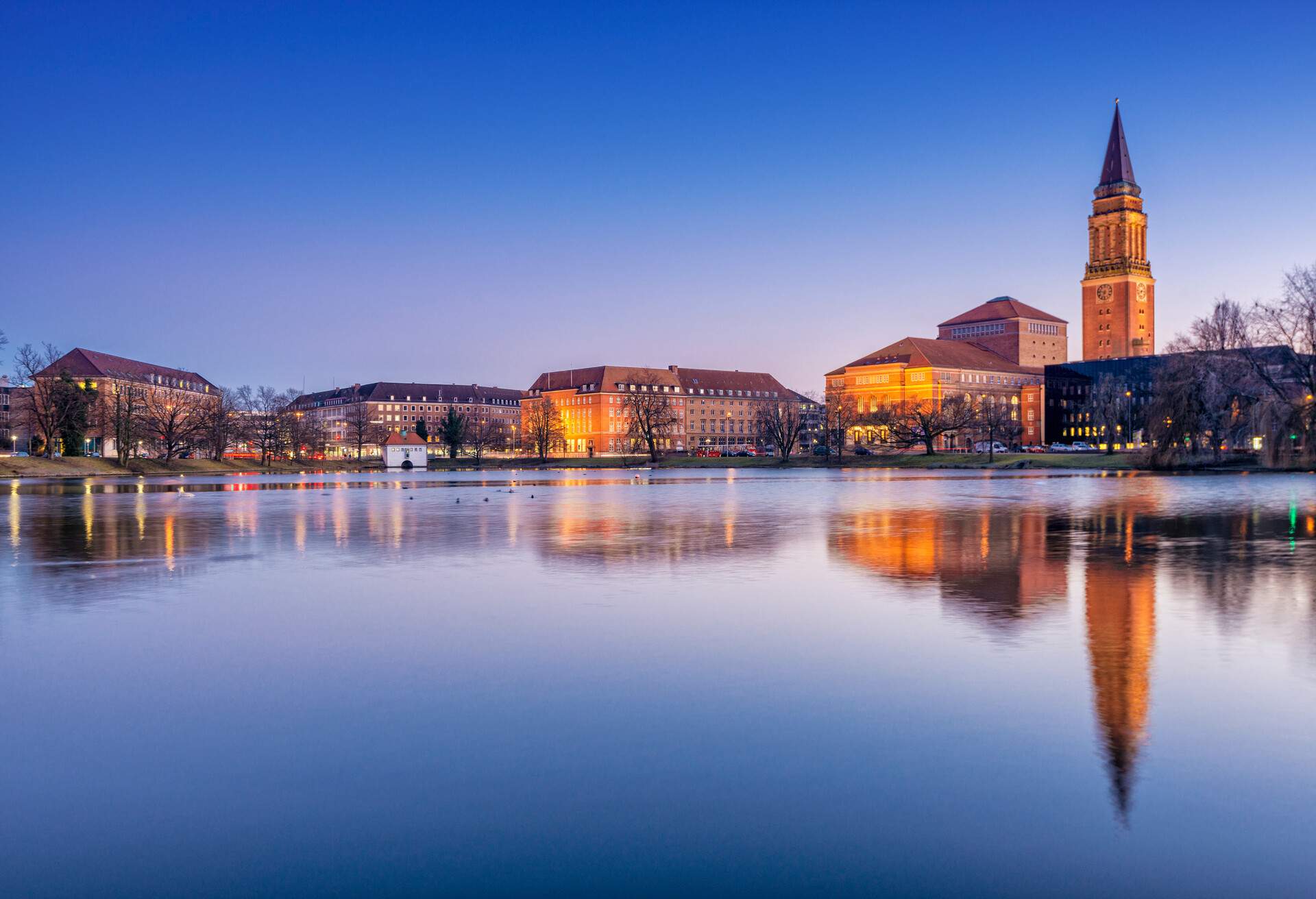 The surface of the lake reflects the illuminated city buildings and clock tower.