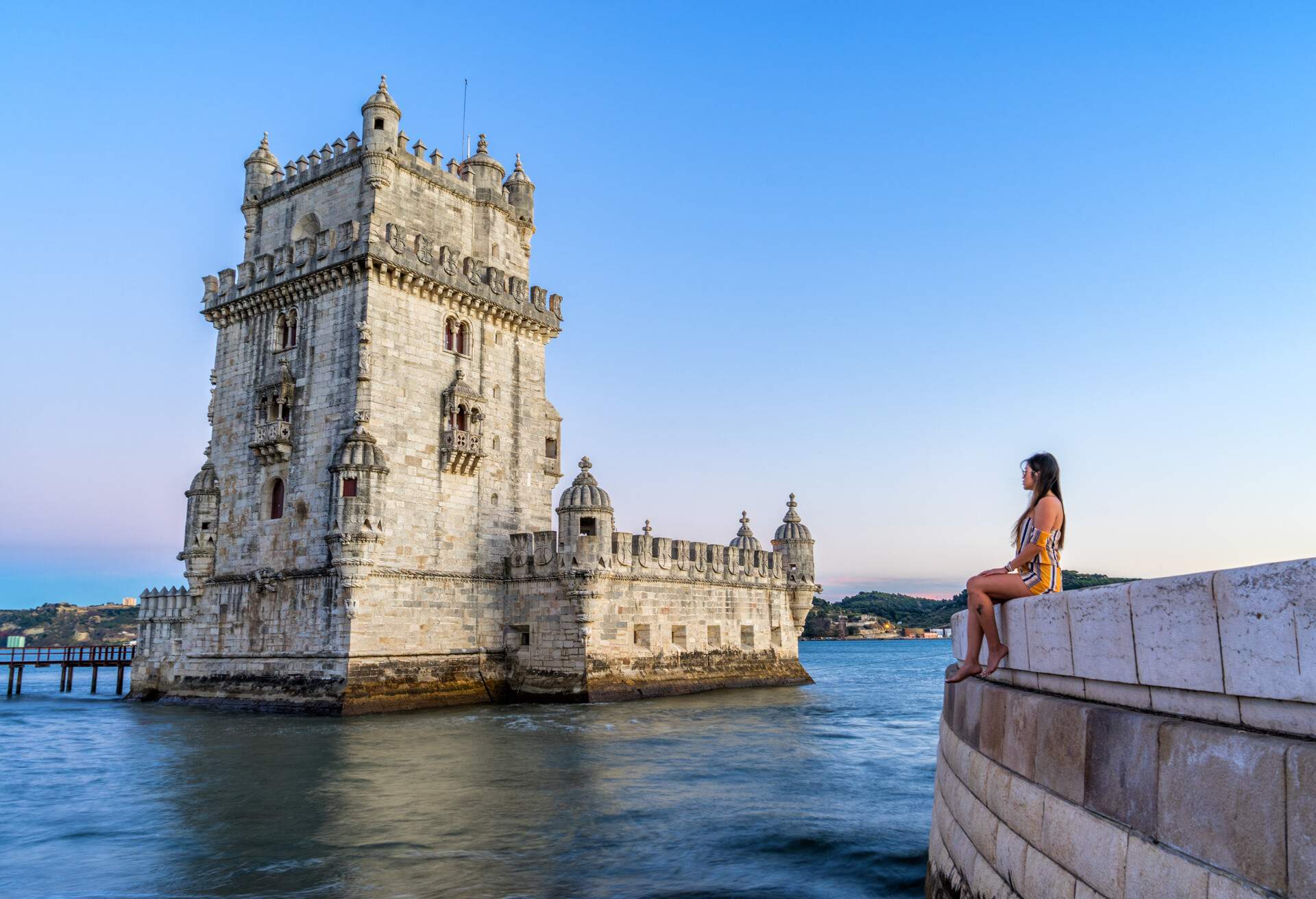 A woman sitting on a stone wall across from the Belém Tower.