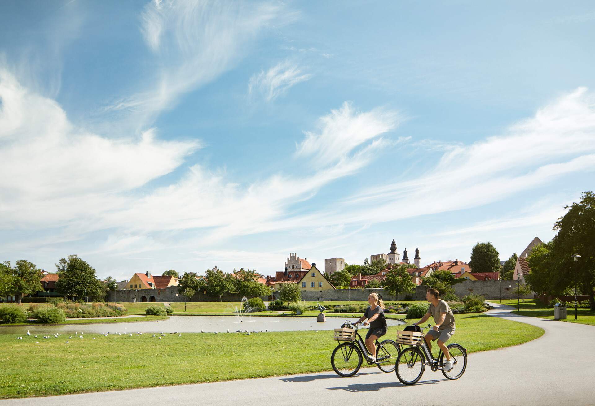 Two cyclists ride down a path in a green park along a pond with a fountain.