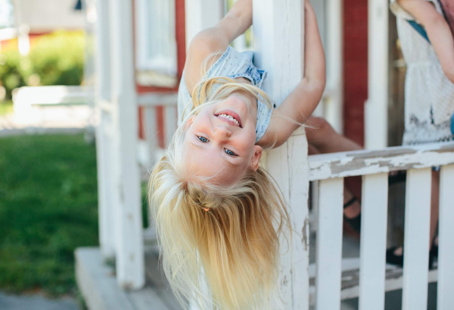 A blonde girl leaning her head upside down on the balcony of a red wooden house 