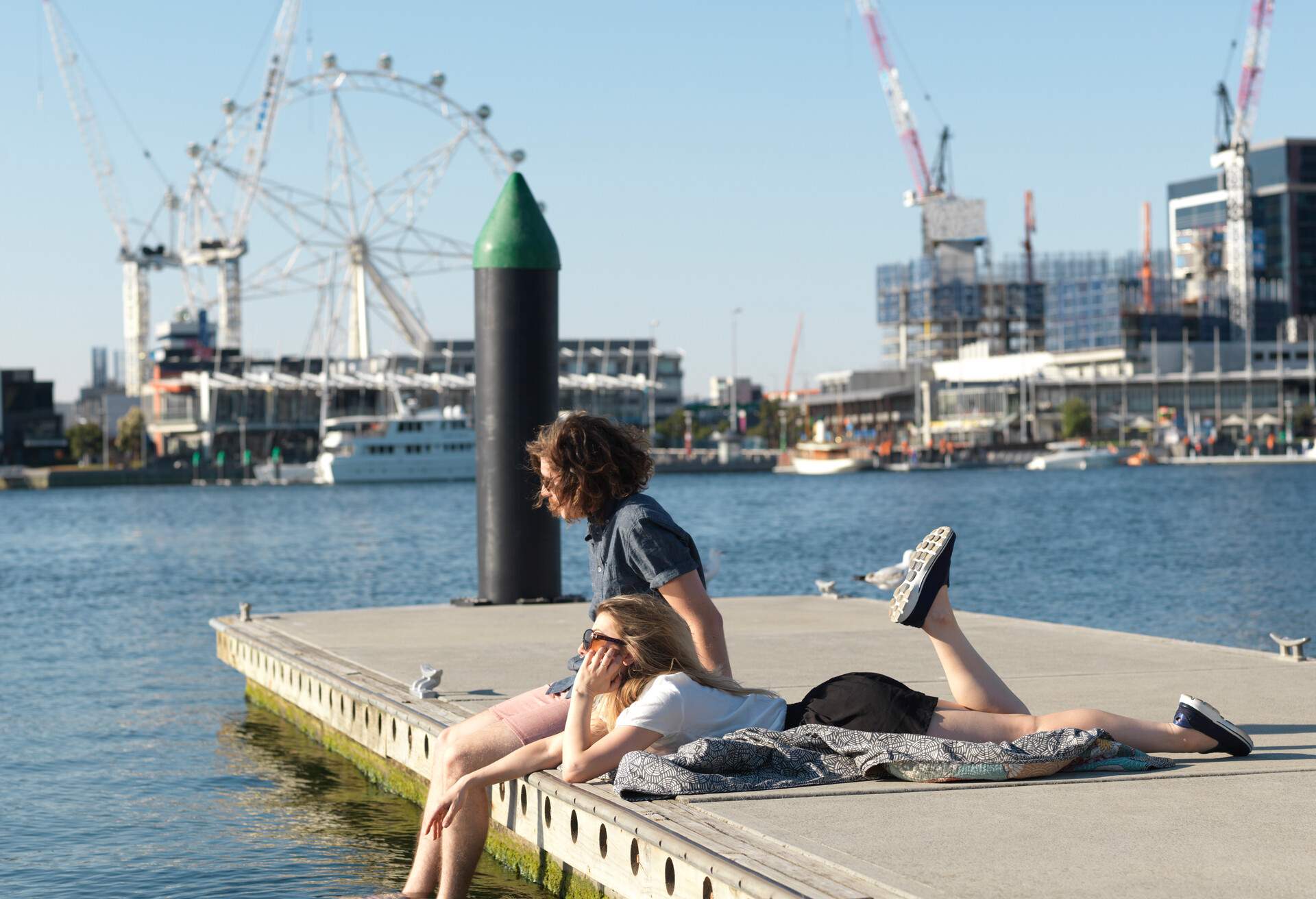 Two friends relaxing on a pier with distant views of a Ferris wheel and construction cranes beside a port.