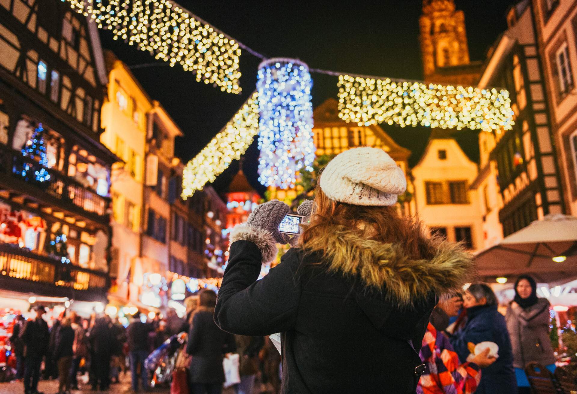 A woman using her camera to capture the scene at a market square with lights.