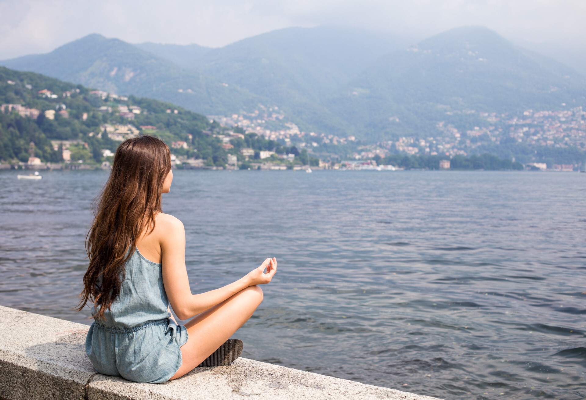 A girl sits serenely by the stone railing, assuming a meditation pose, while a coastal town nestled amidst verdant hills gracefully reflects across the tranquil waters of the lake.