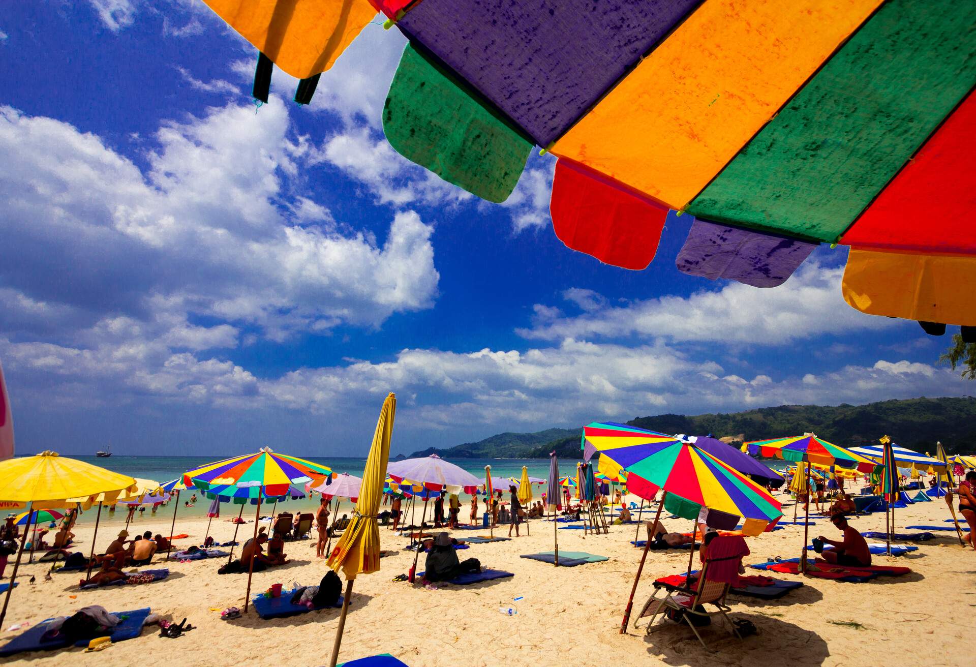 A busy beach full of people with colourful sunbeds and parasols overlooking the sea against the cloudy blue sky.