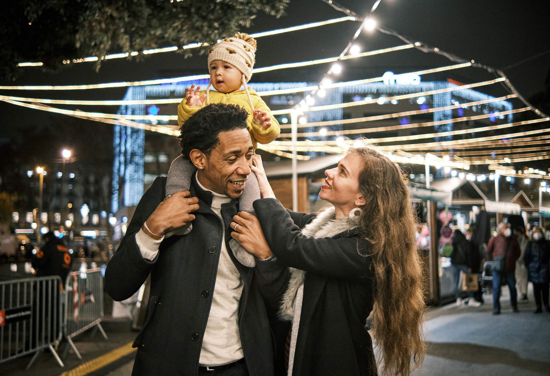 A father with a child on his shoulders and a smiling mother looking at her baby in front of a brightly lit Christmas market.