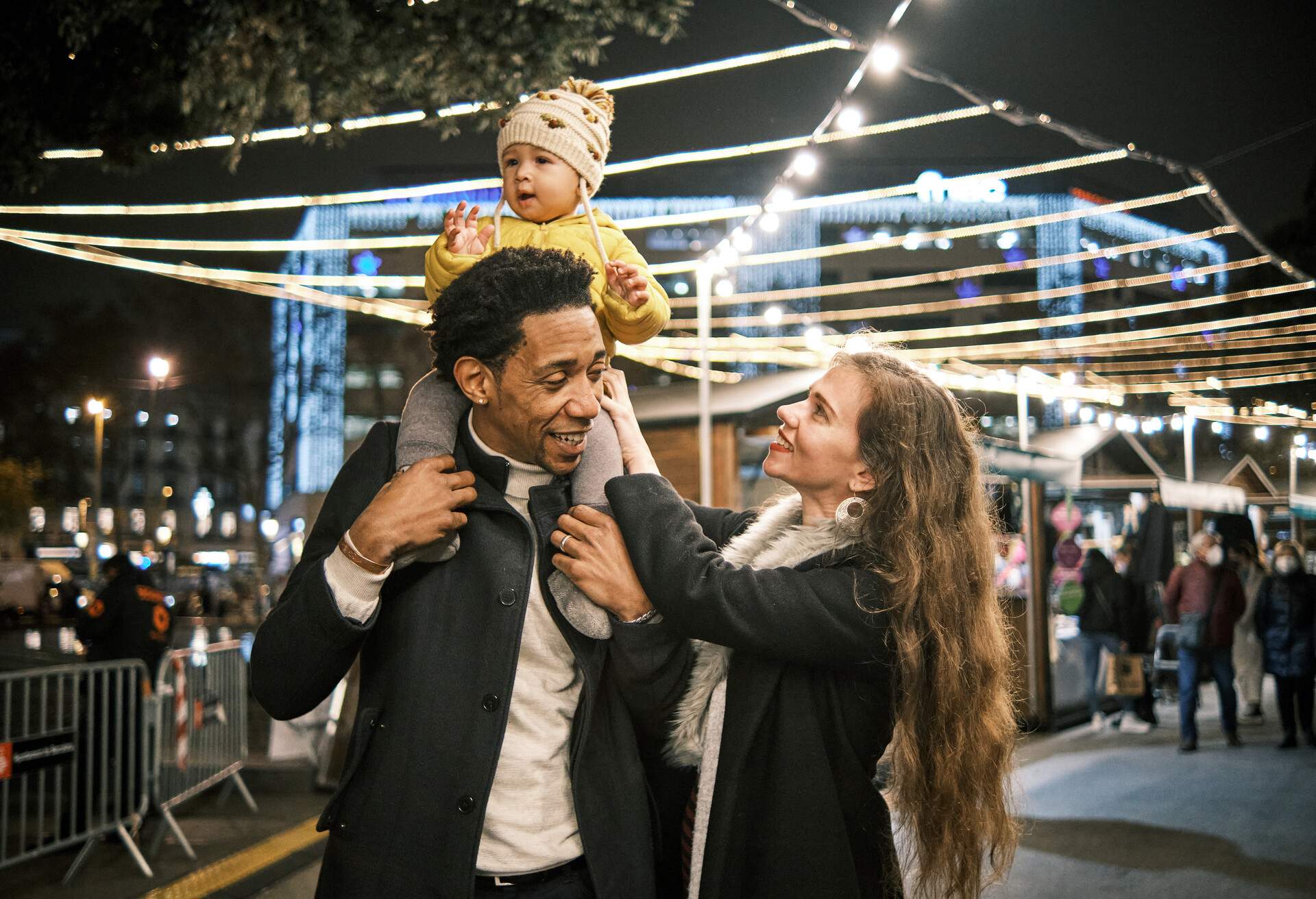 A father with a child on his shoulders and a smiling mother looking at her baby in front of a brightly lit Christmas market.