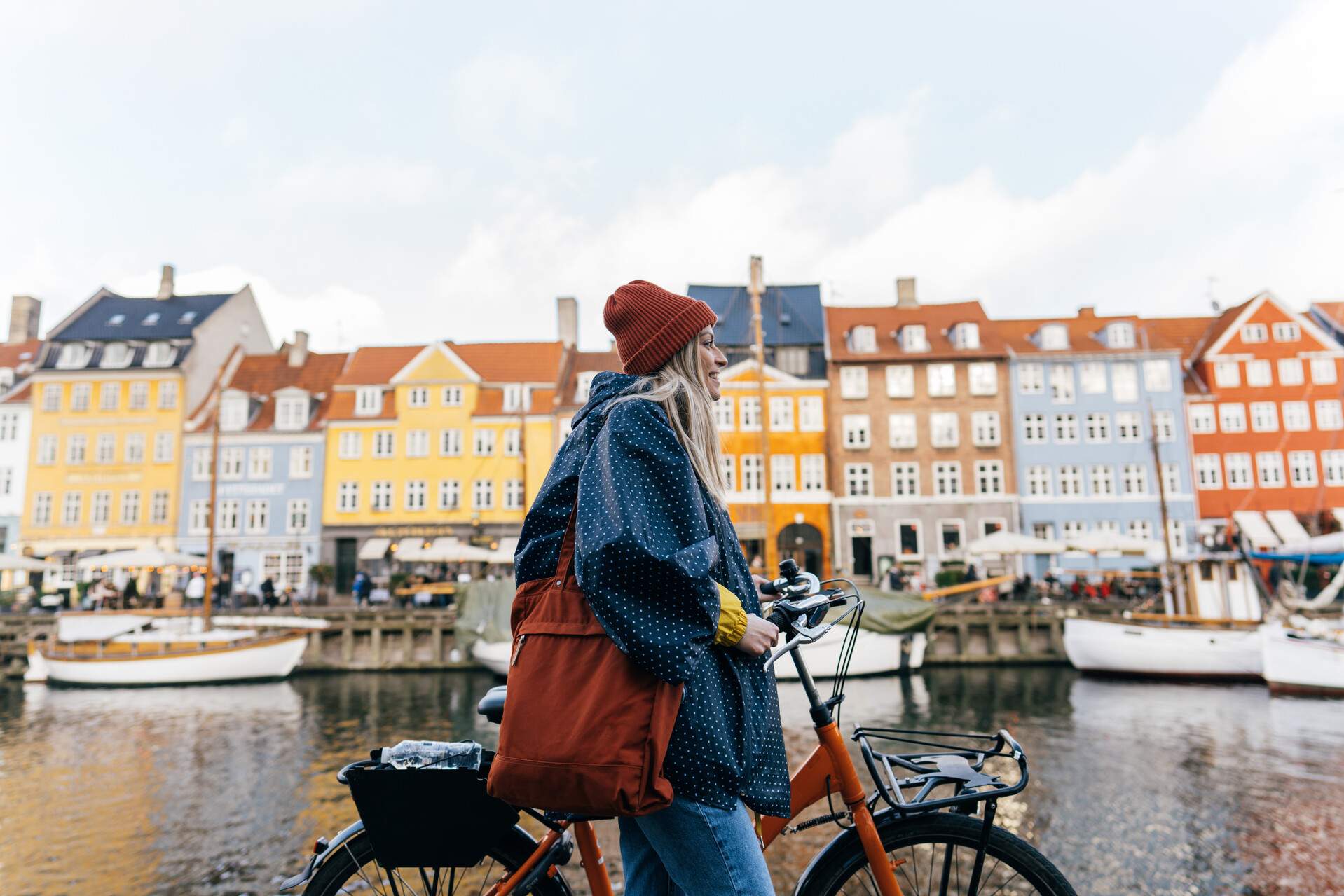 A woman in casual clothes stands beside a bicycle with a backdrop of a river canal along a row of colourful buildings.