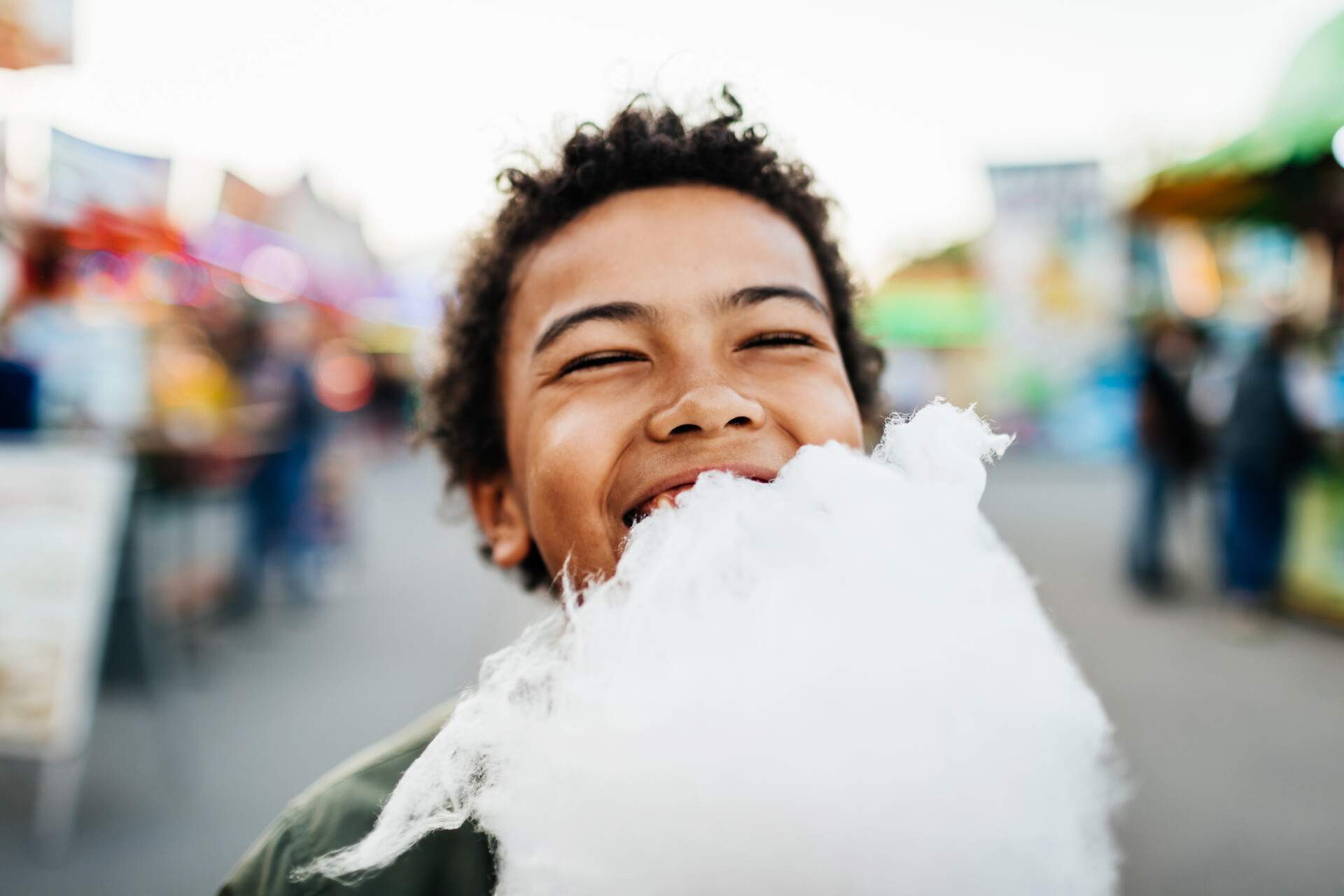 A kid with dark hair is smiling and eating a cotton candy