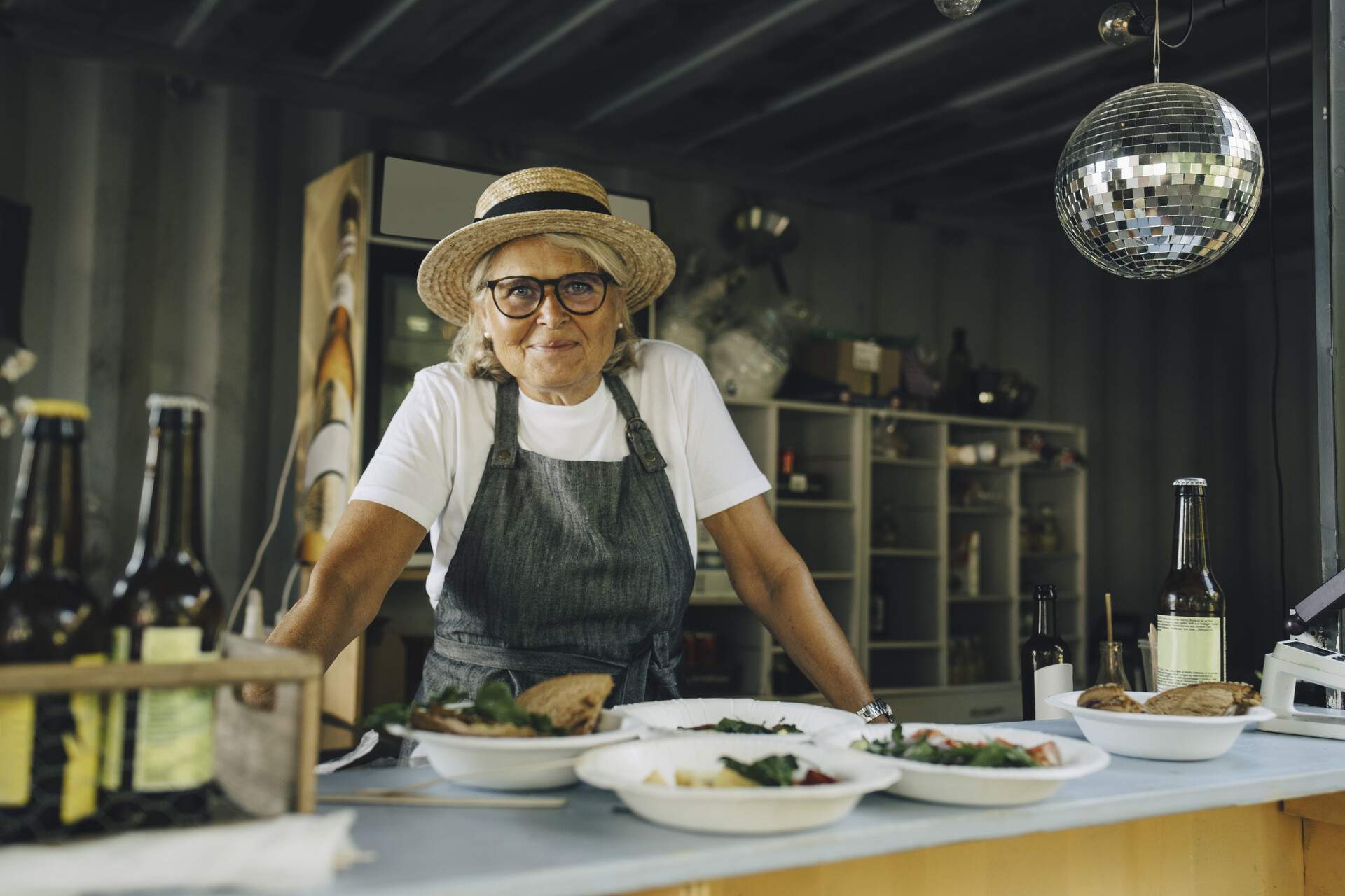 An elderly woman in a straw hat and apron stands beside a restaurant counter full of food.