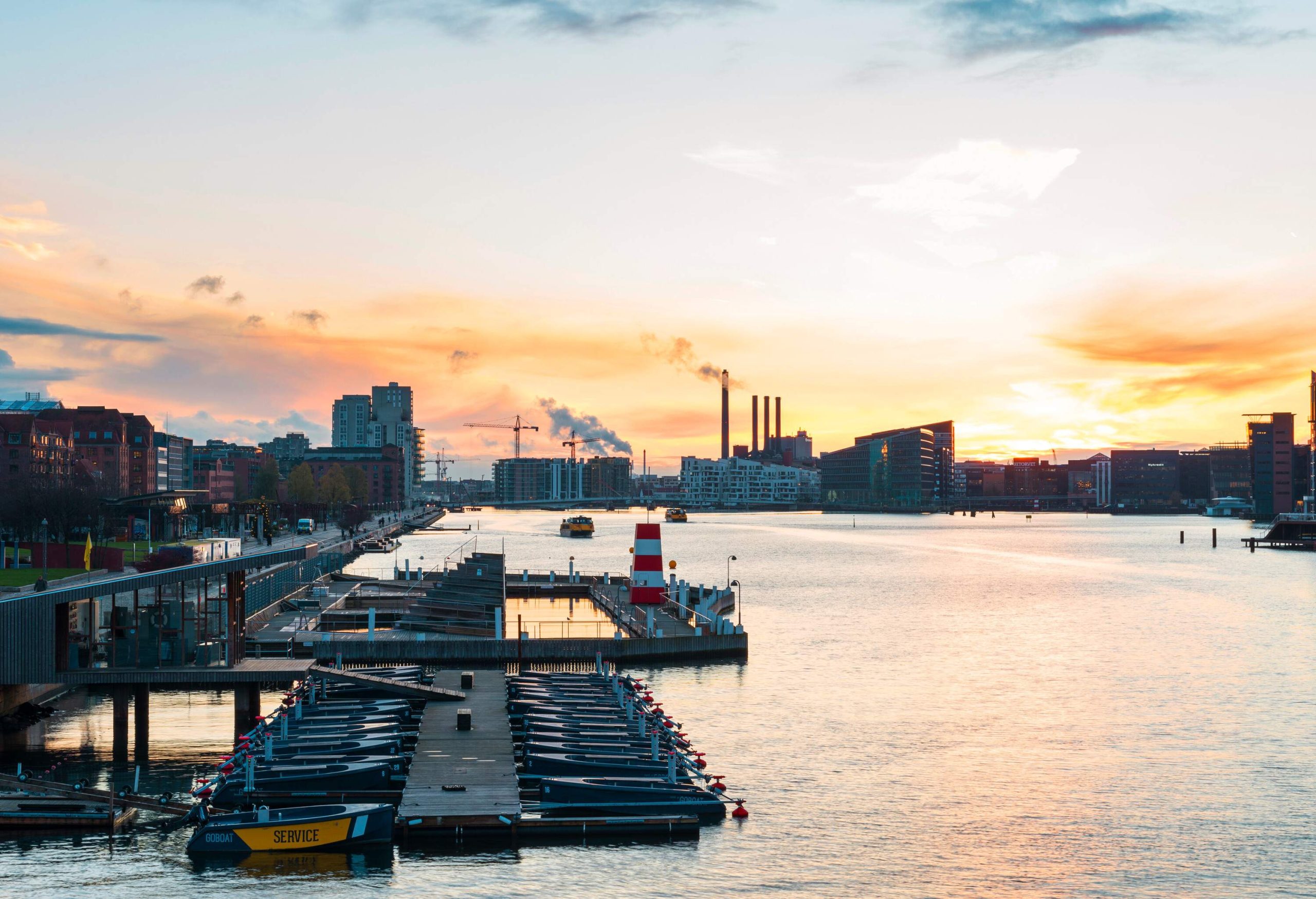 A harbour with anchored boats and distant views of a cityscape's silhouette during sunset.