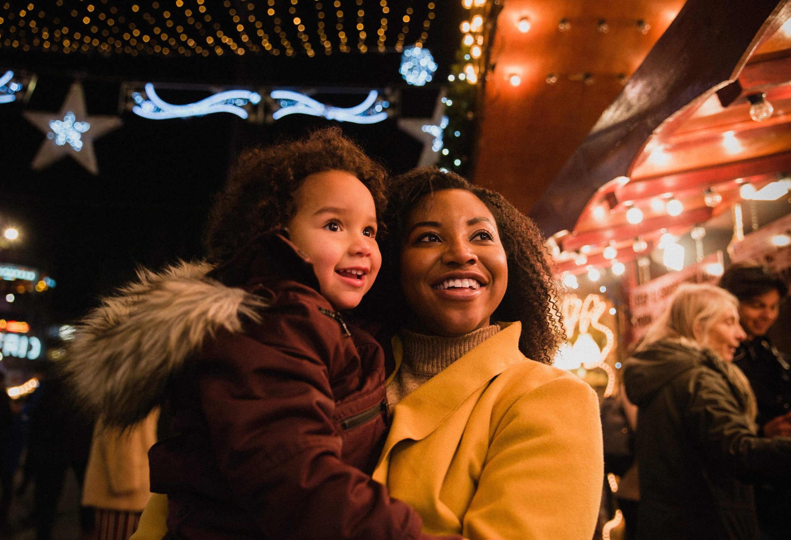 Smiling woman holding her child with a look of awe against the background of colourfully illuminated lights.