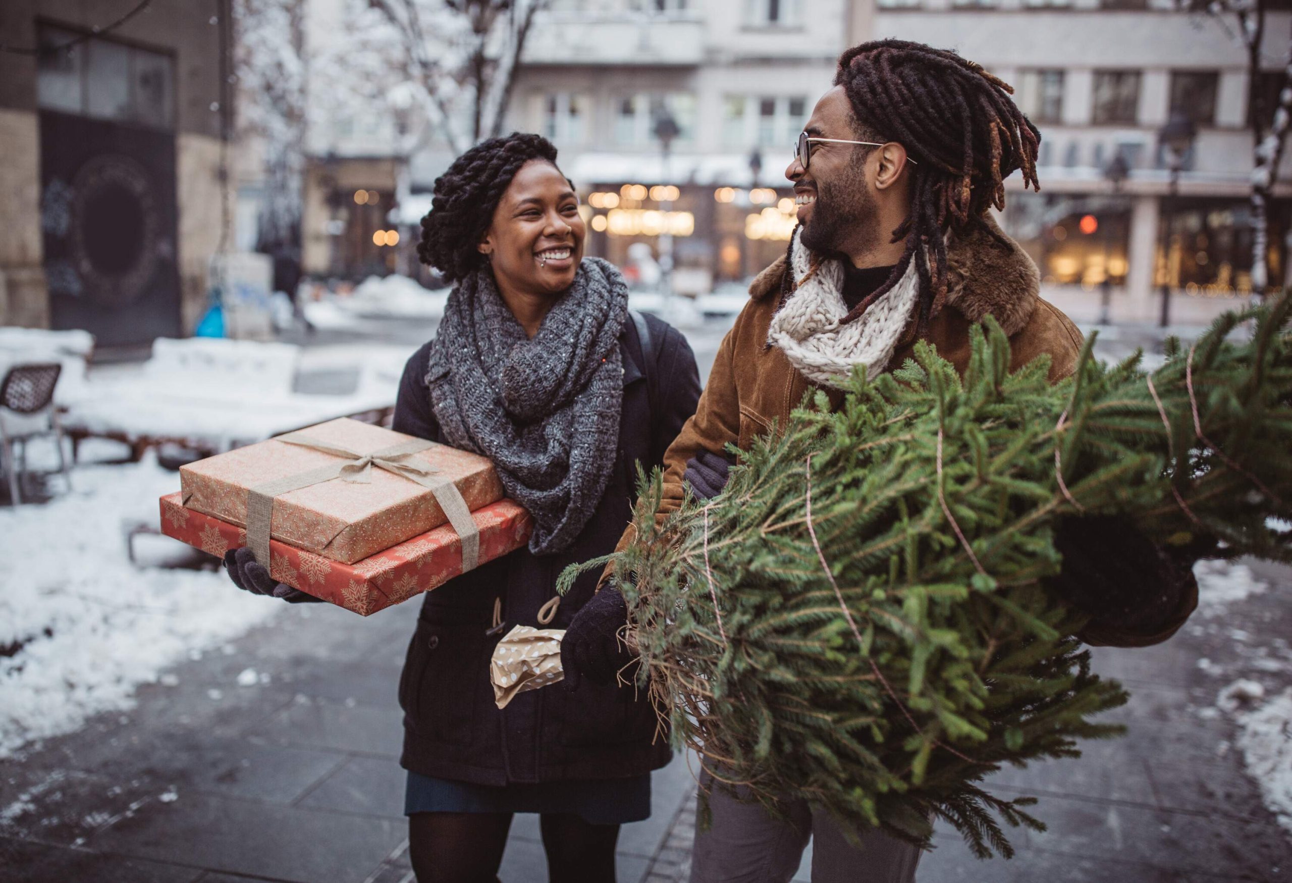 A couple in winter clothes smile at each other as they carry wrapped gifts and Christmas tree.