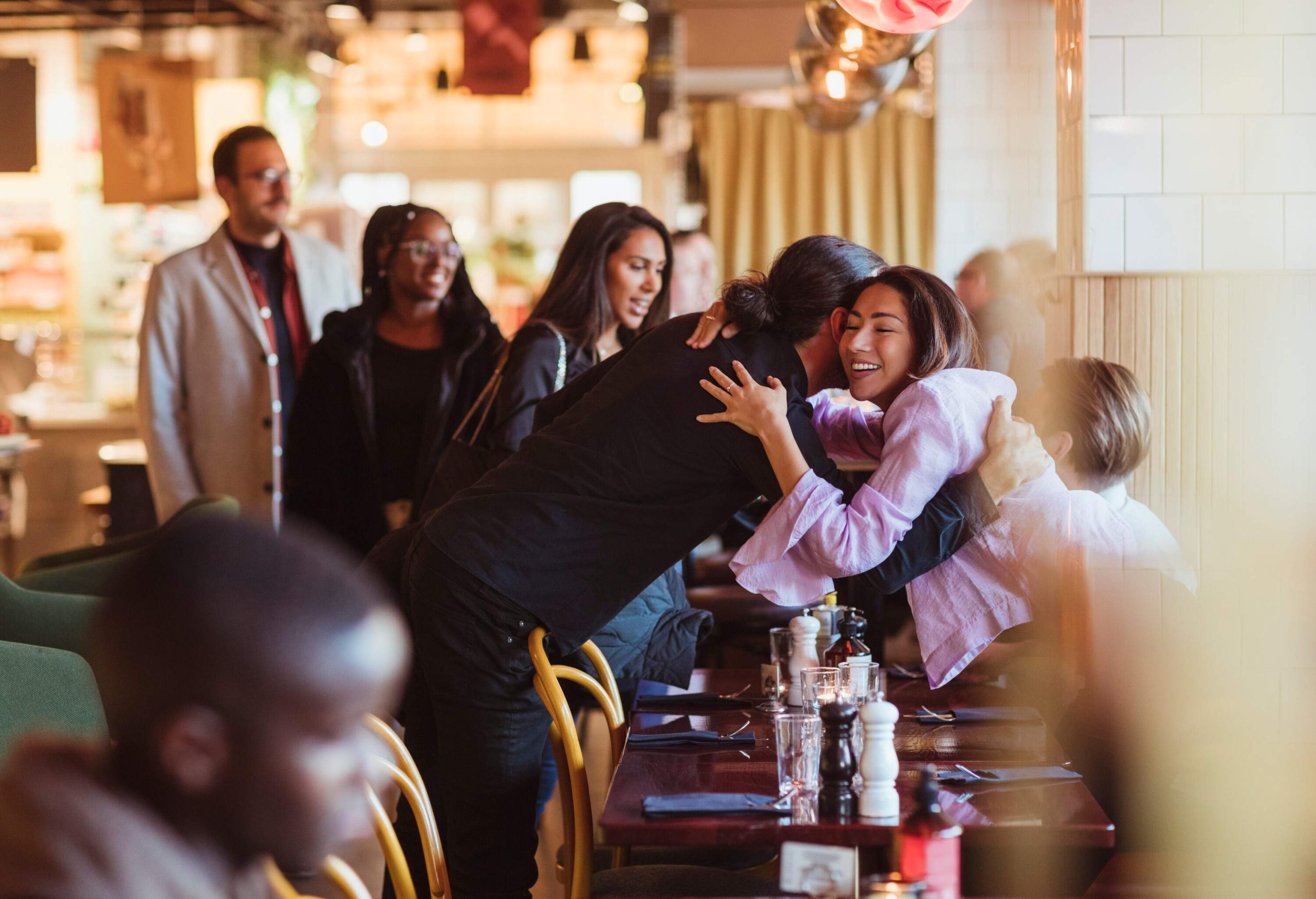 A circle of male and females friends hugs upon arriving at the restaurant.