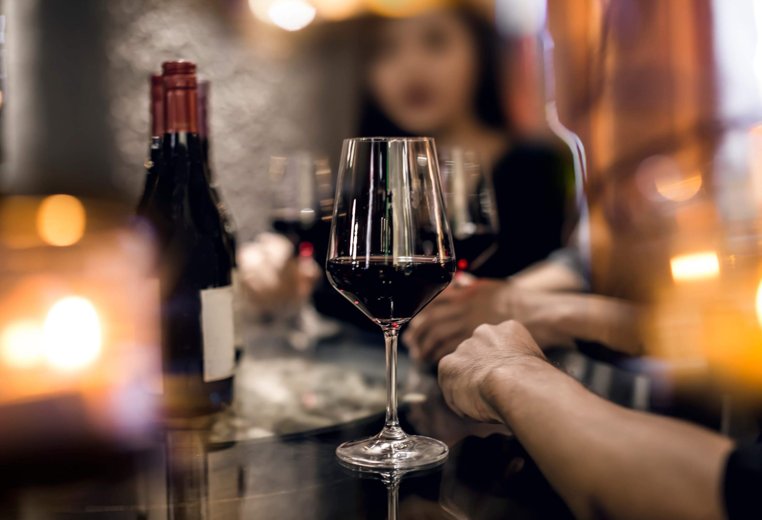 A red wine glass is placed on a table, surrounded by a group of friends in the background, creating a blurred effect.