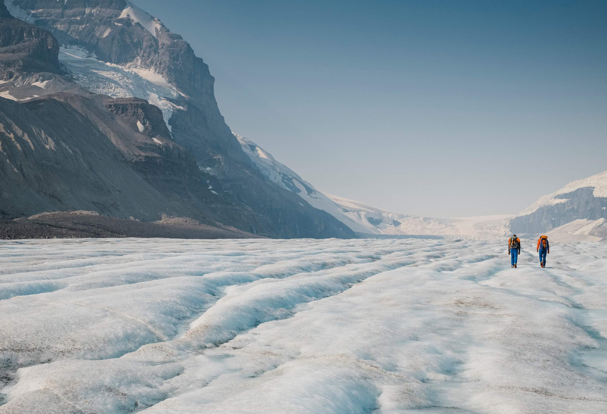 Two people walk on a vast glacier beneath the rocky mountains.