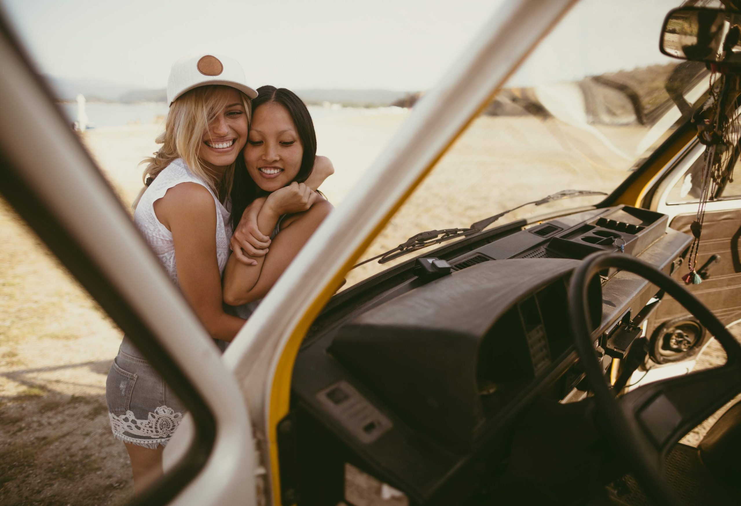Two happy women hug in front of a vehicle parked by the shore.
