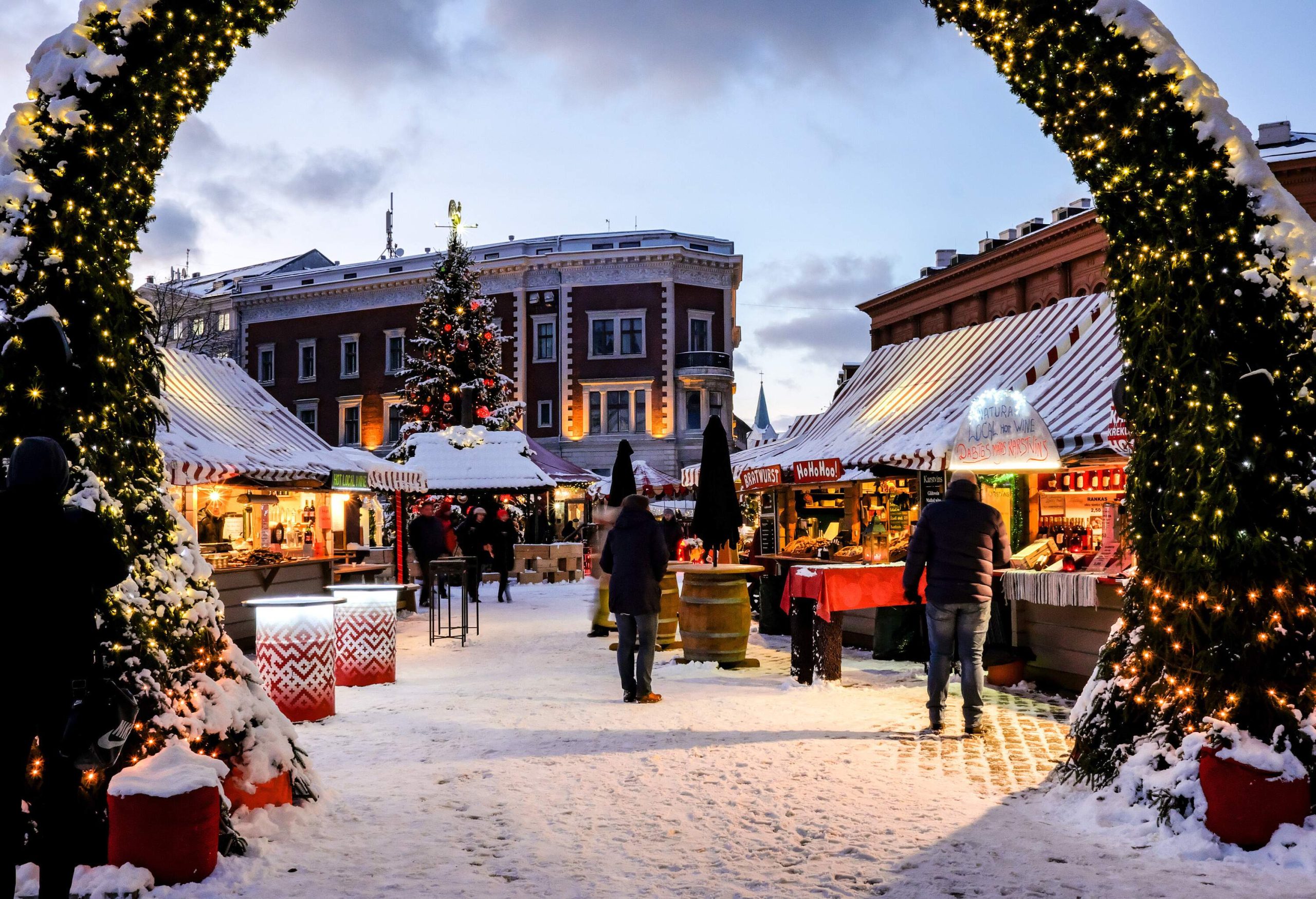 An entrance archway to a snow-covered Christmas market decorated with lights and garlands.