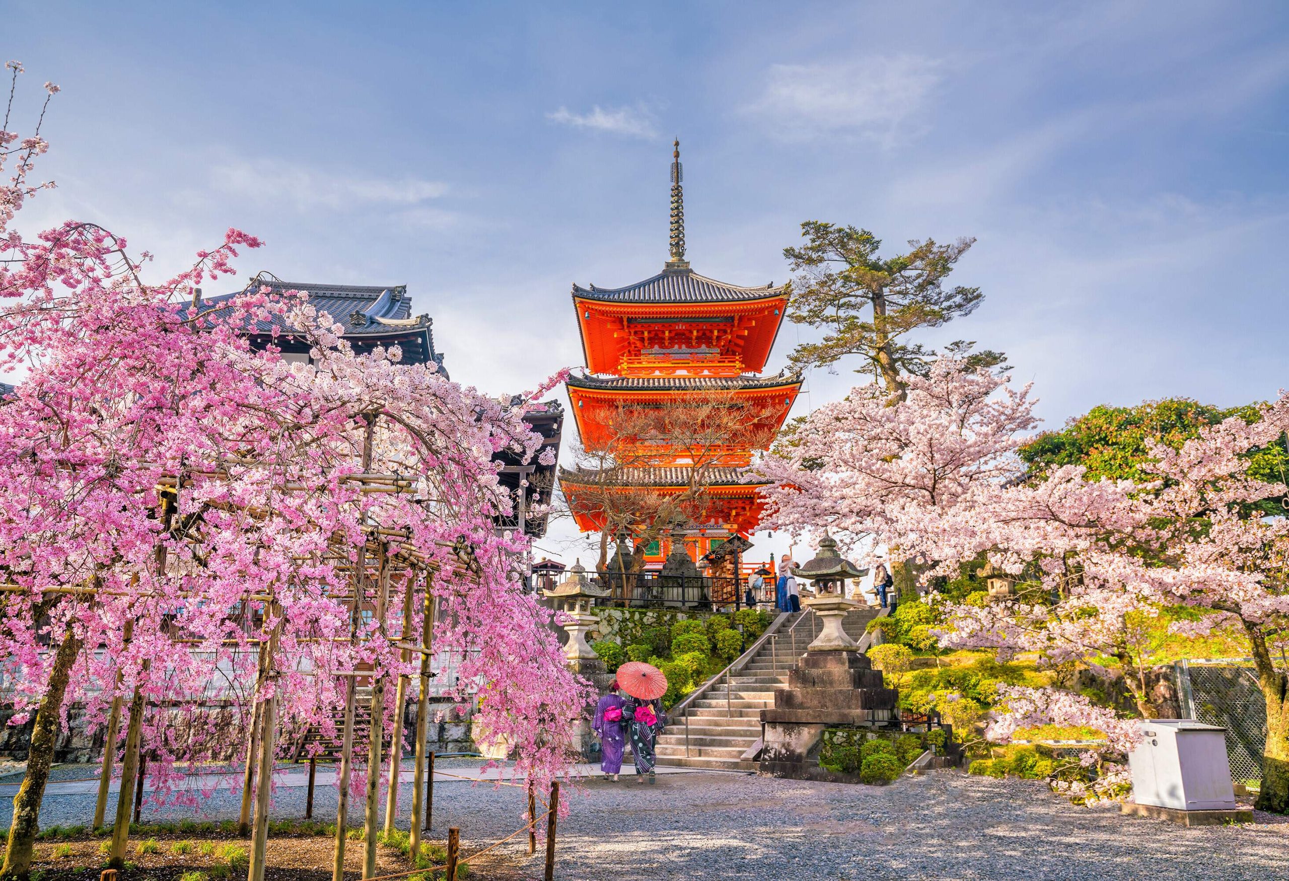 A three-tier Japanese pagoda-like temple with a bright orange facade perched atop a hill surrounded by beautiful cherry blossoms.