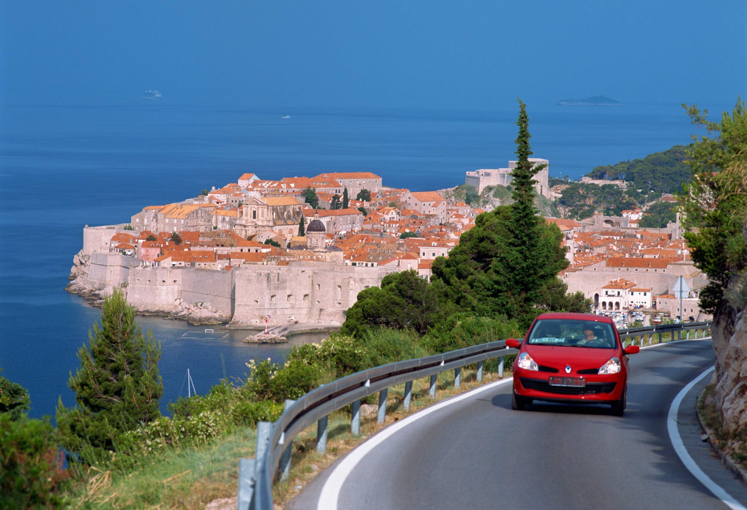 A vibrant red car traverses a winding road atop a hill, offering breathtaking views of a walled coastal city below.