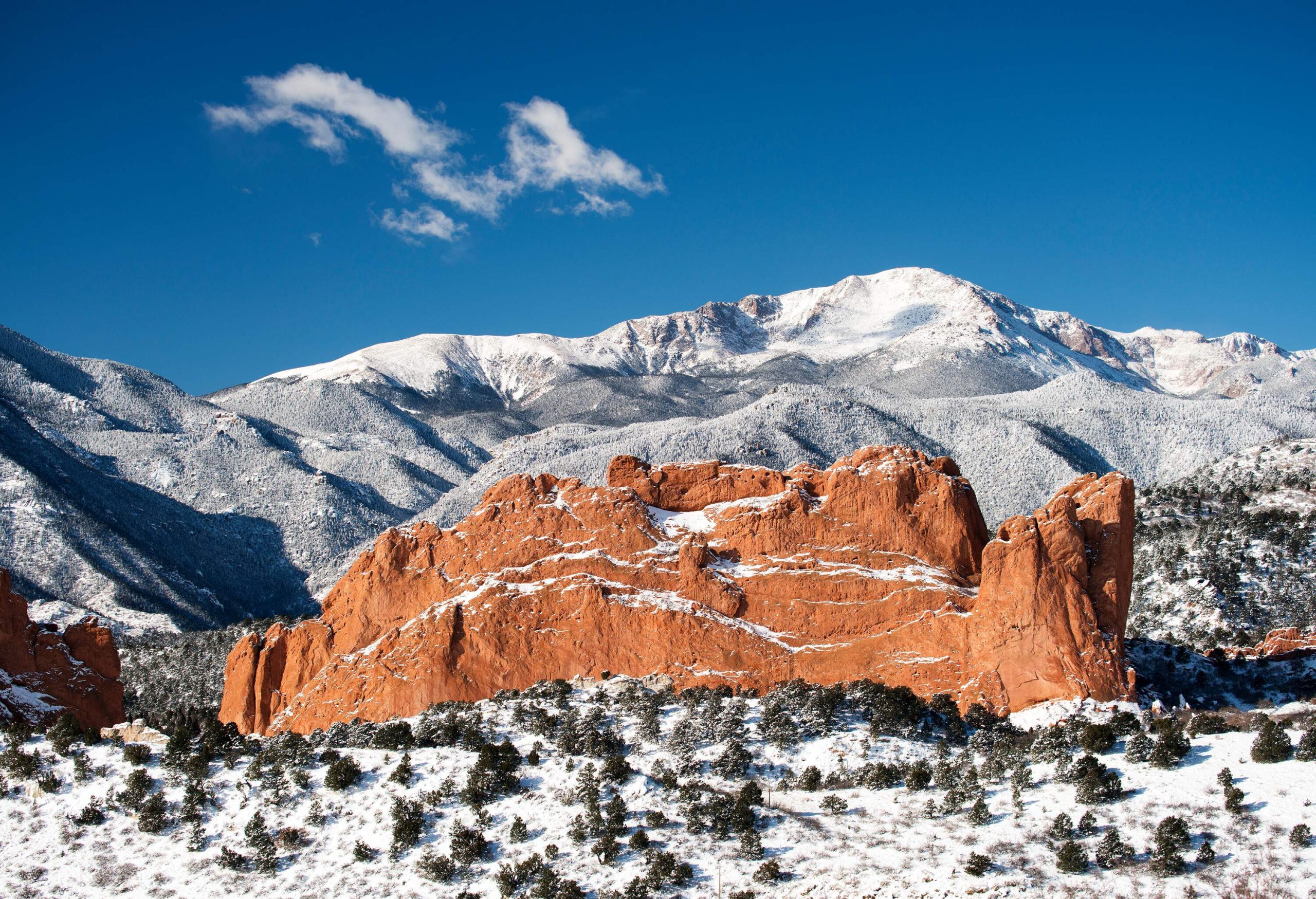 A spectacular sandstone rock formation emerges from the ground at the foot of a snow-capped rocky mountain.
