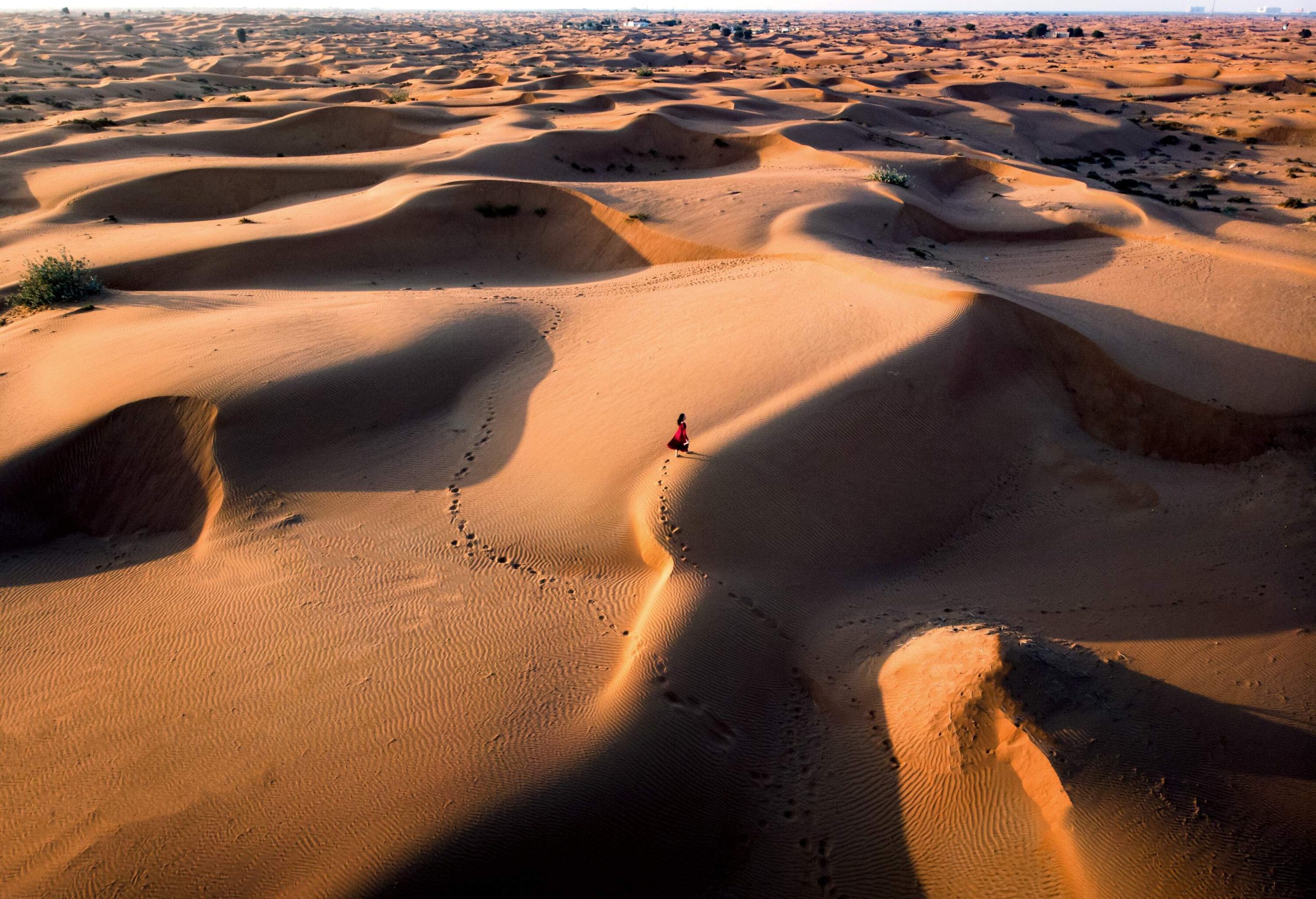 A woman in a dress walks in the middle of the desert, leaving her footprints on the dunes.