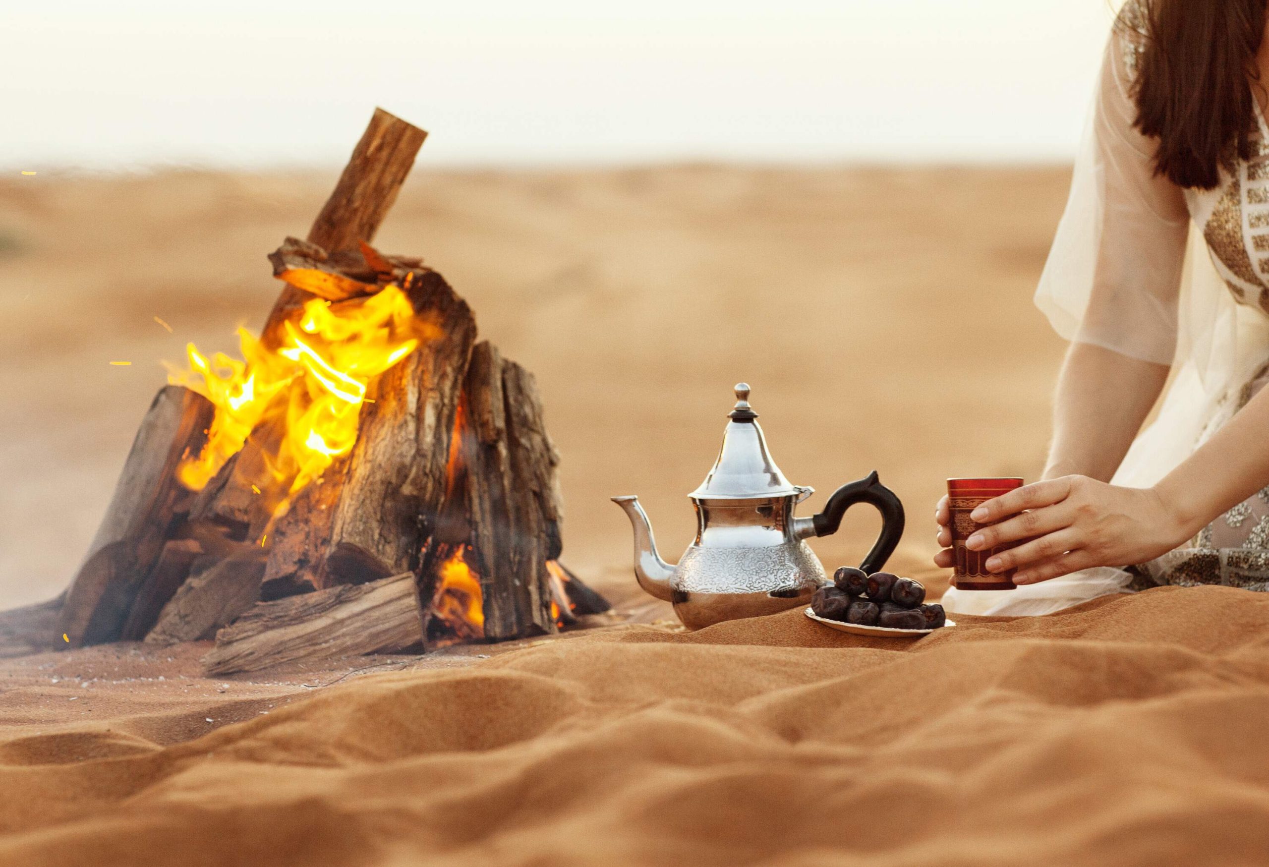 A woman holding a cup sitting next to an Arabic teapot and a small plate of dates by a campfire on a sand dune.
