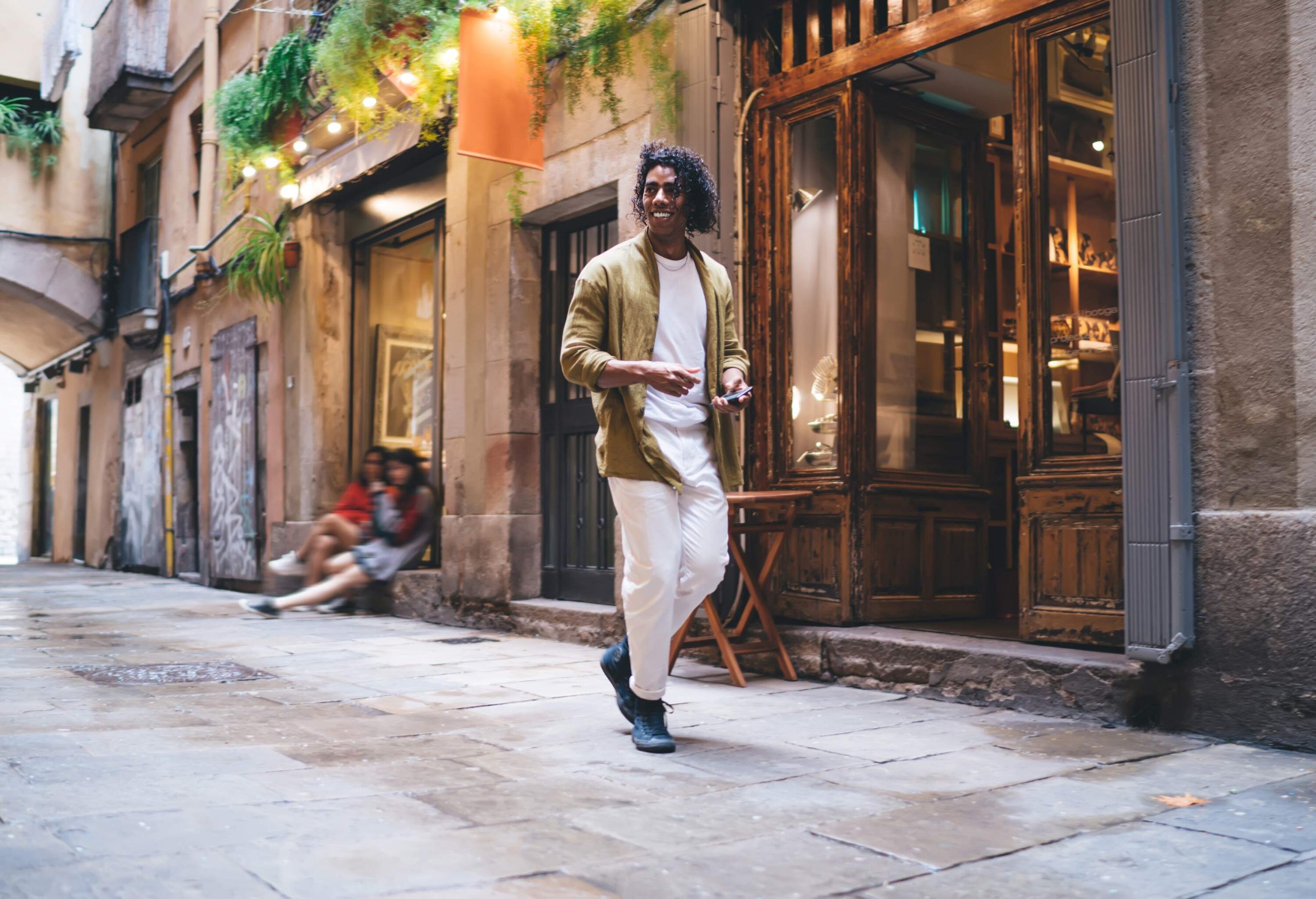 A man in a white outfit and brown blazer happily walking down a cobbled street in front of classic building stalls.