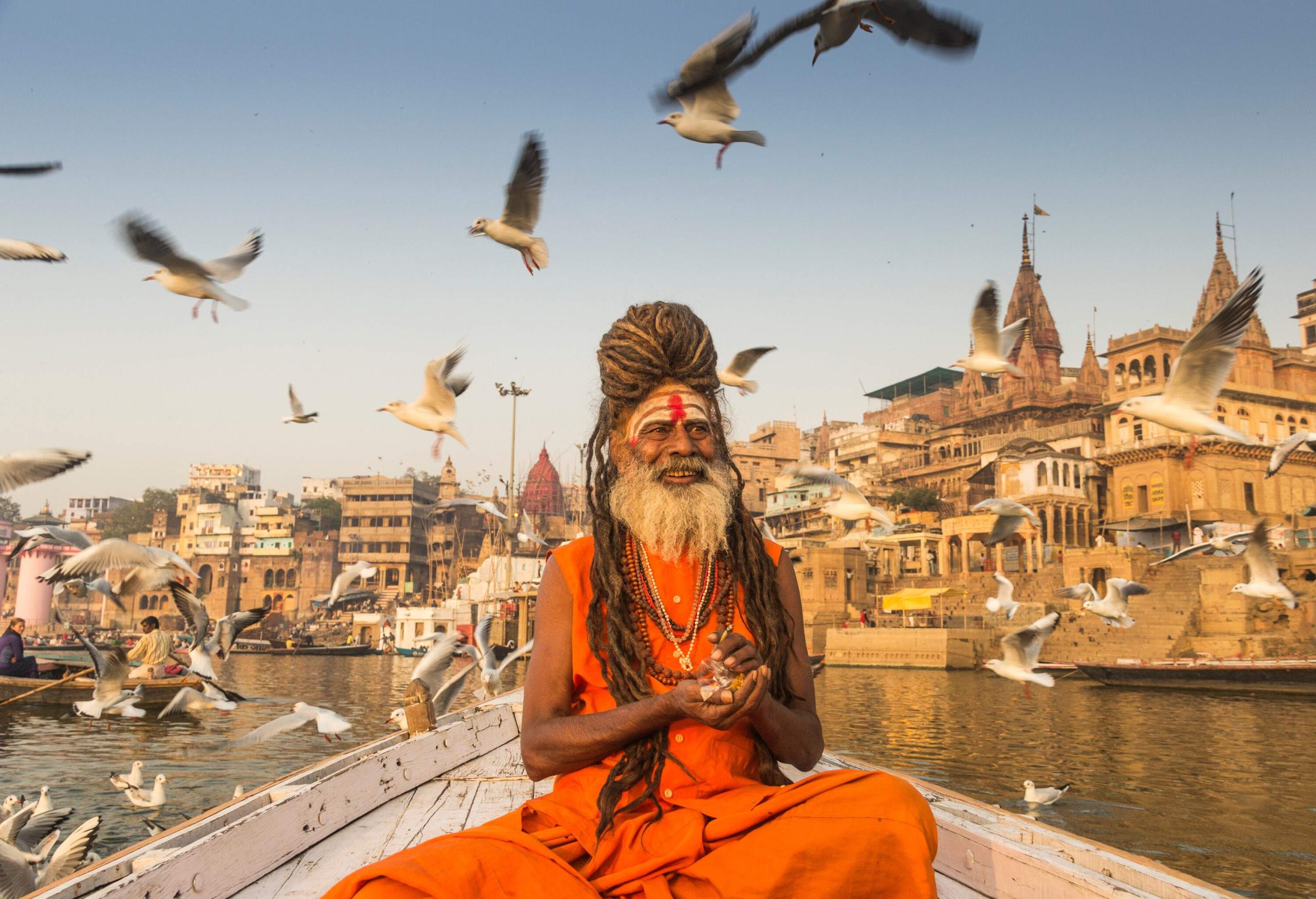 A sadhu wearing an orange saffron sits at the boat's bow, surrounded by flying birds while sailing in the river.